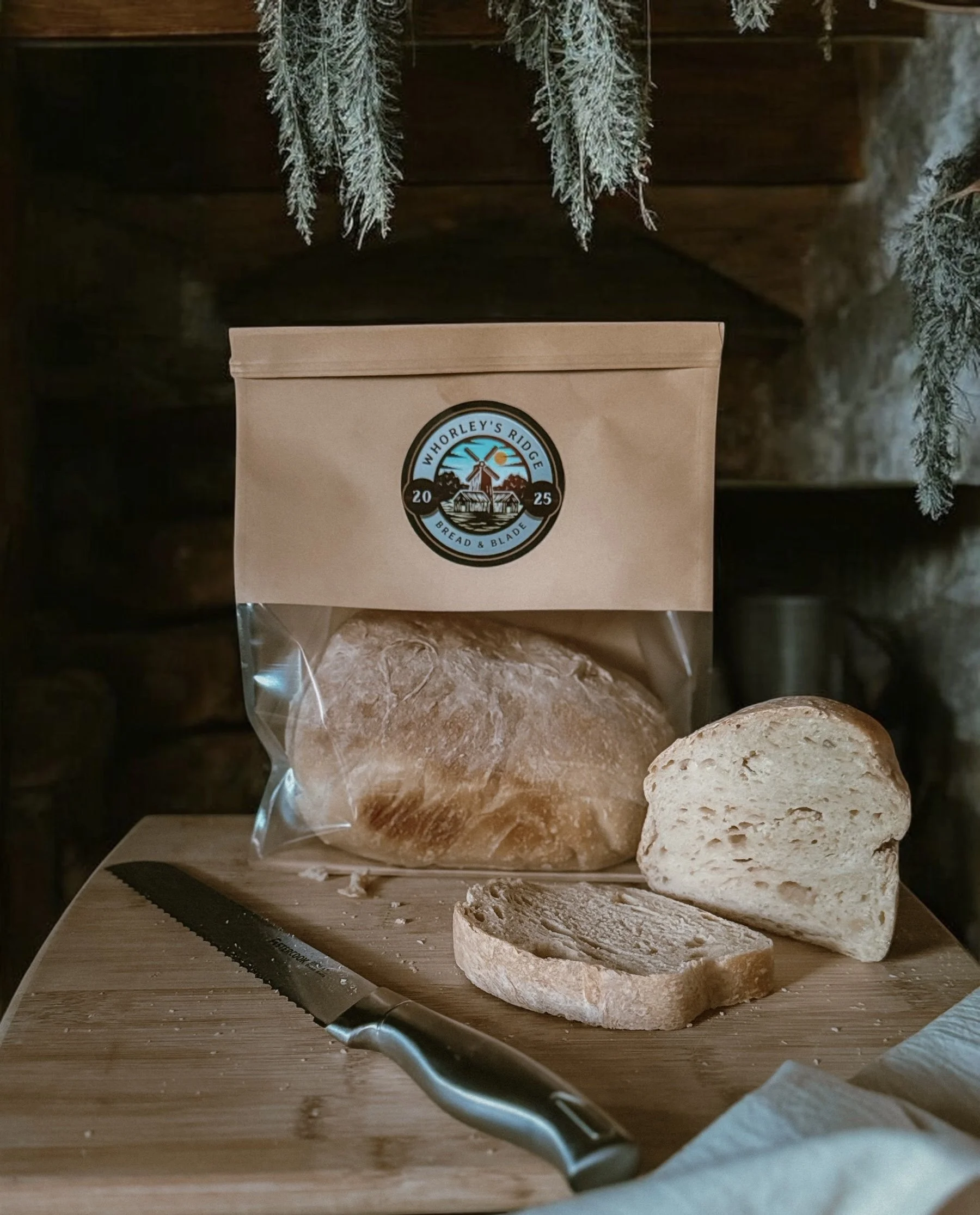 A loaf of bread, sliced bread, and a bread knife on a wooden cutting board, with a bag of bread from Whorley's Ridge Bakery in the background, set against a rustic wooden and stone background.