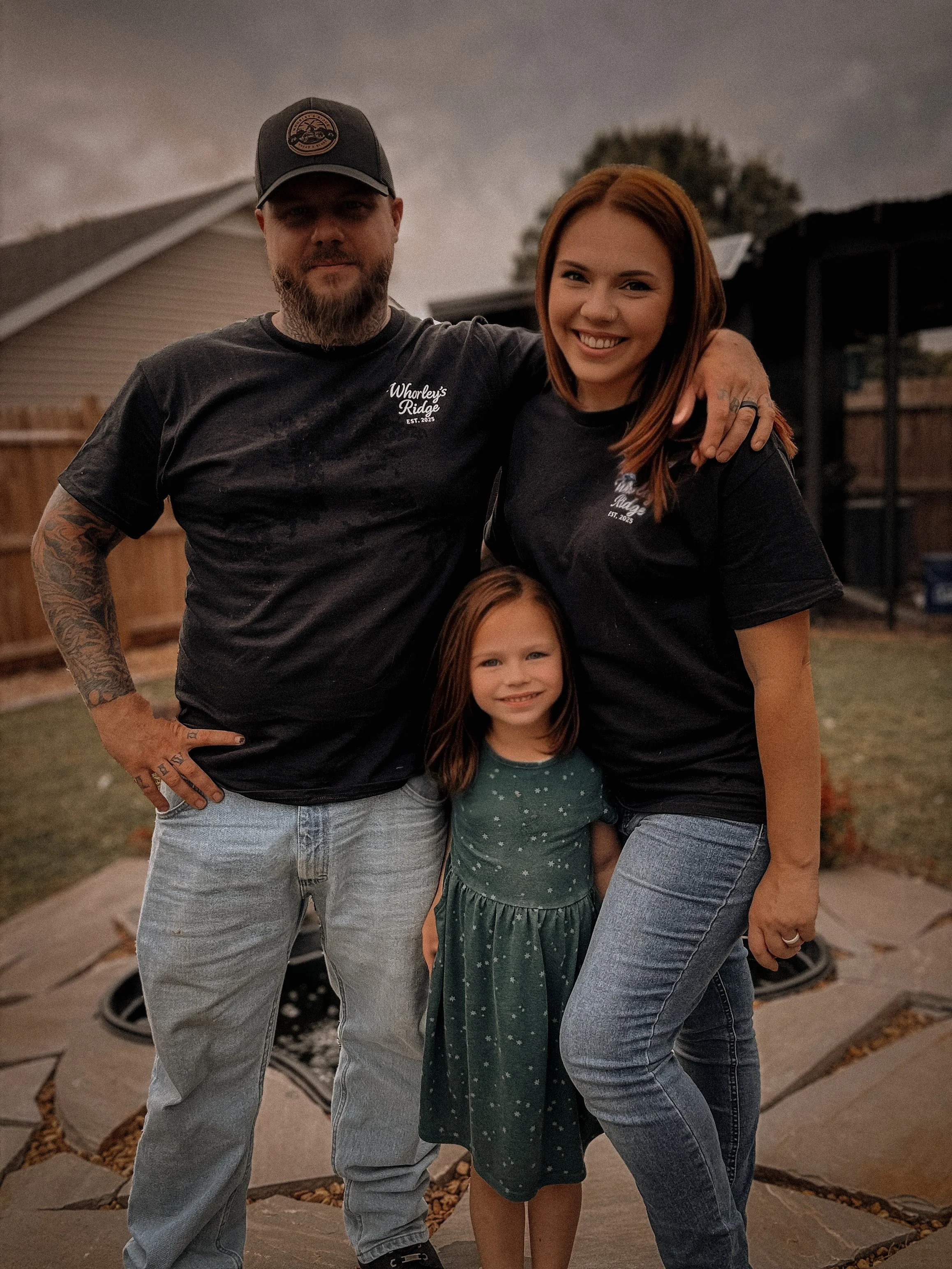 A family of three standing outdoors in a backyard during evening, smiling at the camera. The father has a beard, tattoos, and wears a black baseball cap and T-shirt. The mother has red hair and wears a black T-shirt. Their young daughter has red hair and wears a green dress with white stars.
