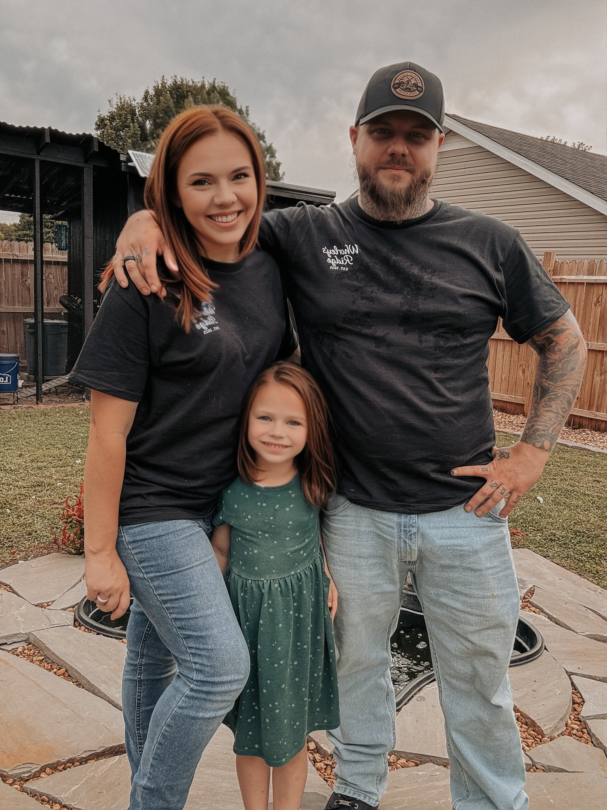 A family of four posing outdoors, with a woman, a man, and a young girl in front of a backyard with a wooden fence, a small pond, and a house.