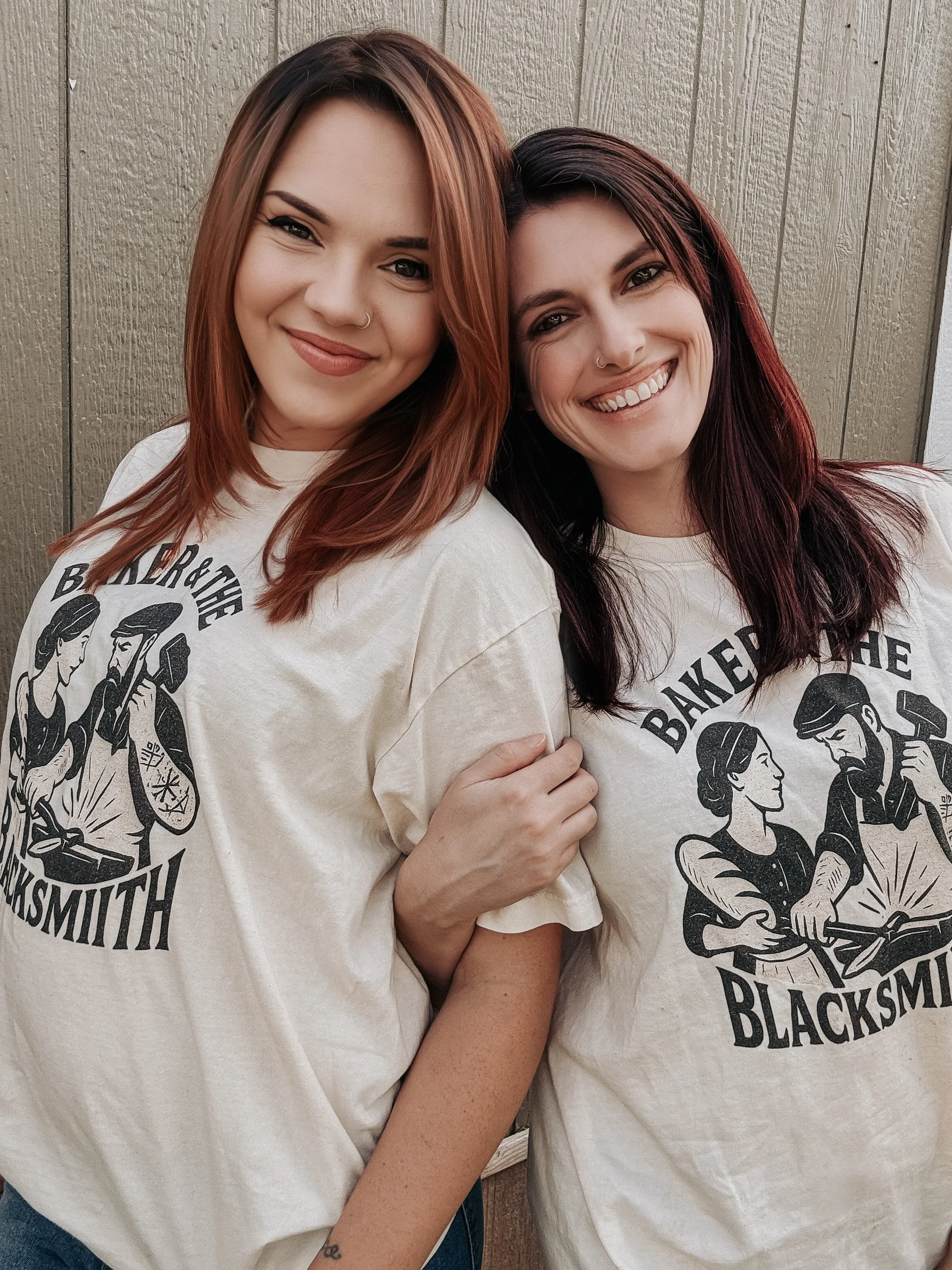 Two women smiling with their heads touching, wearing white T-shirts with a black graphic design and text that reads 'Bake her the Blacksmith' against a wooden background.