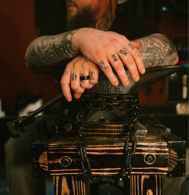Close-up of a tattooed person's hands with rings, resting on a DJ controller with chains, in a dimened-lit setting.