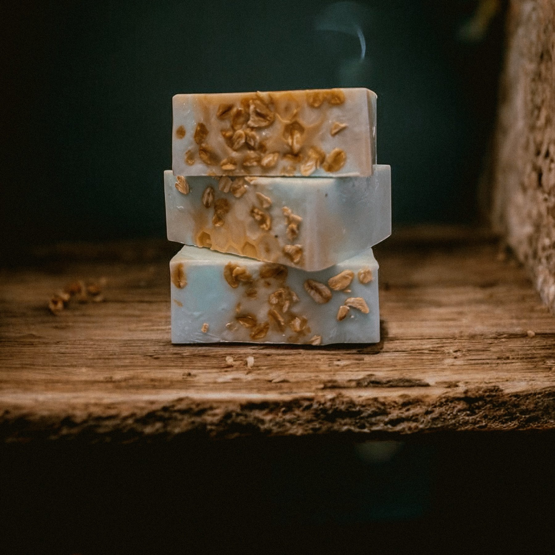 Three stacked bars of handmade soap with oats on top, placed on a rustic wooden surface, with a dark background.