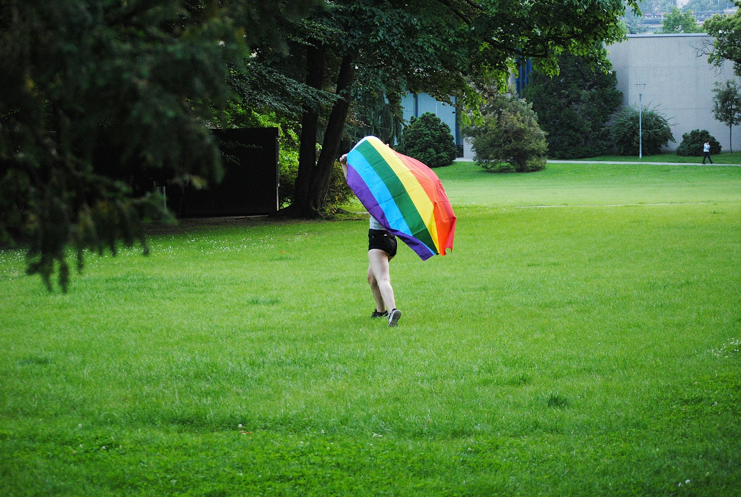 Person holding a rainbow-colored umbrella walking across a grassy park with trees and some buildings in the background.