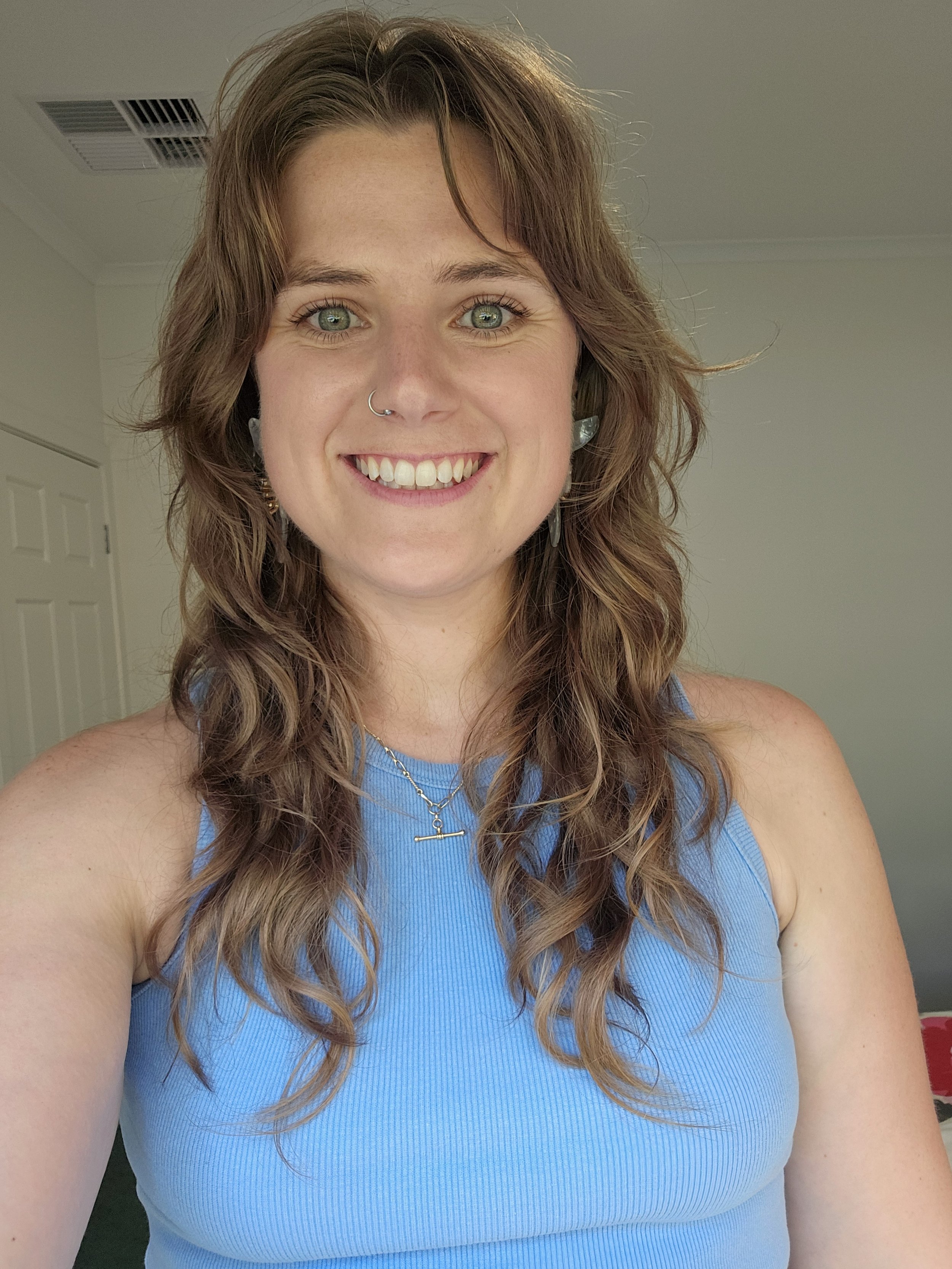 Close-up portrait of Katelyn Gray women's health naturopath with long, curly reddish-brown hair, wearing a blue sleeveless top, earrings, and a necklace, standing in a room with beige walls and a white door in the background smiling