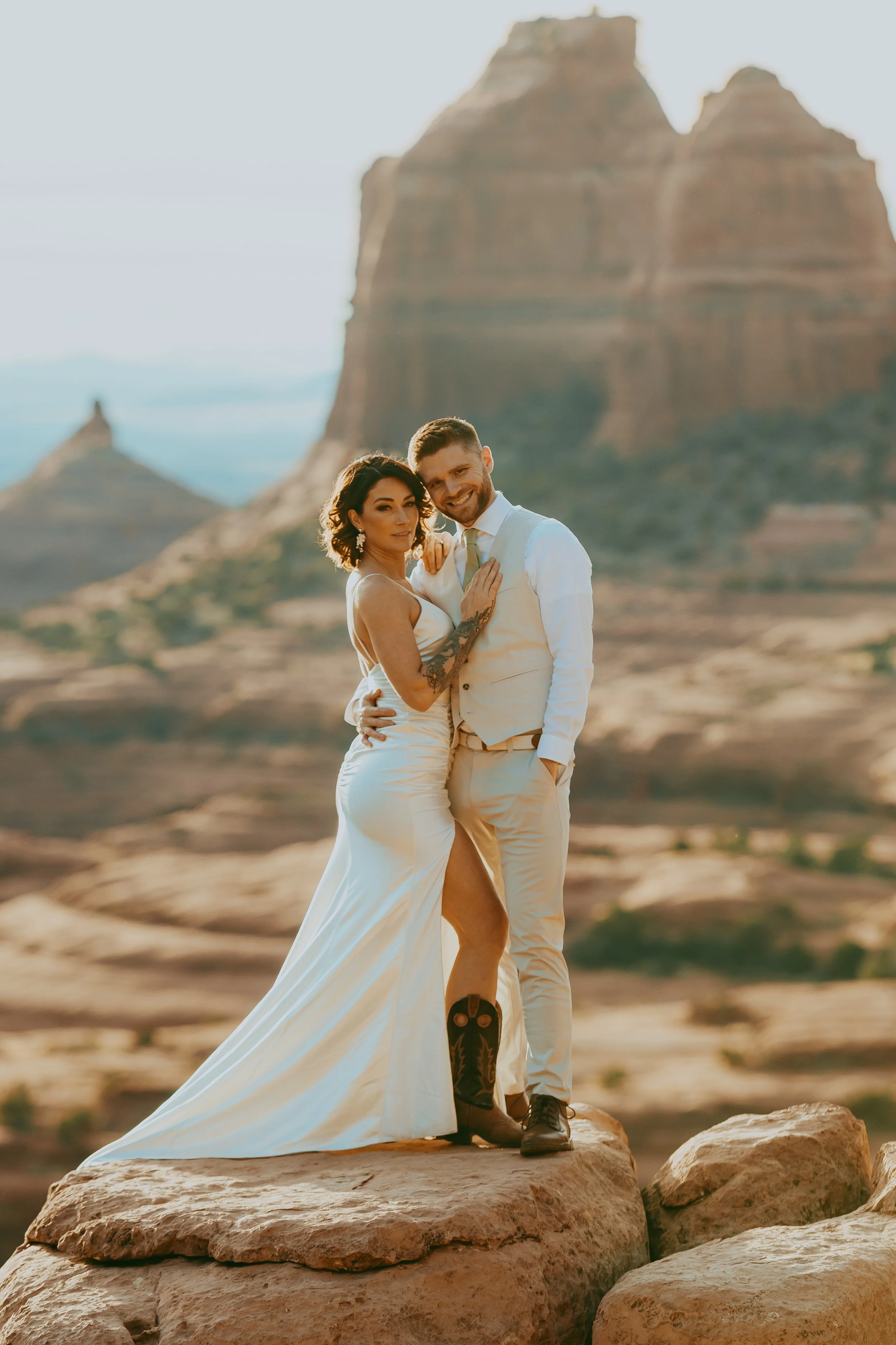 A newlywed couple stands on rocks in a desert landscape with rock formations in the background, smiling and embracing during sunset.