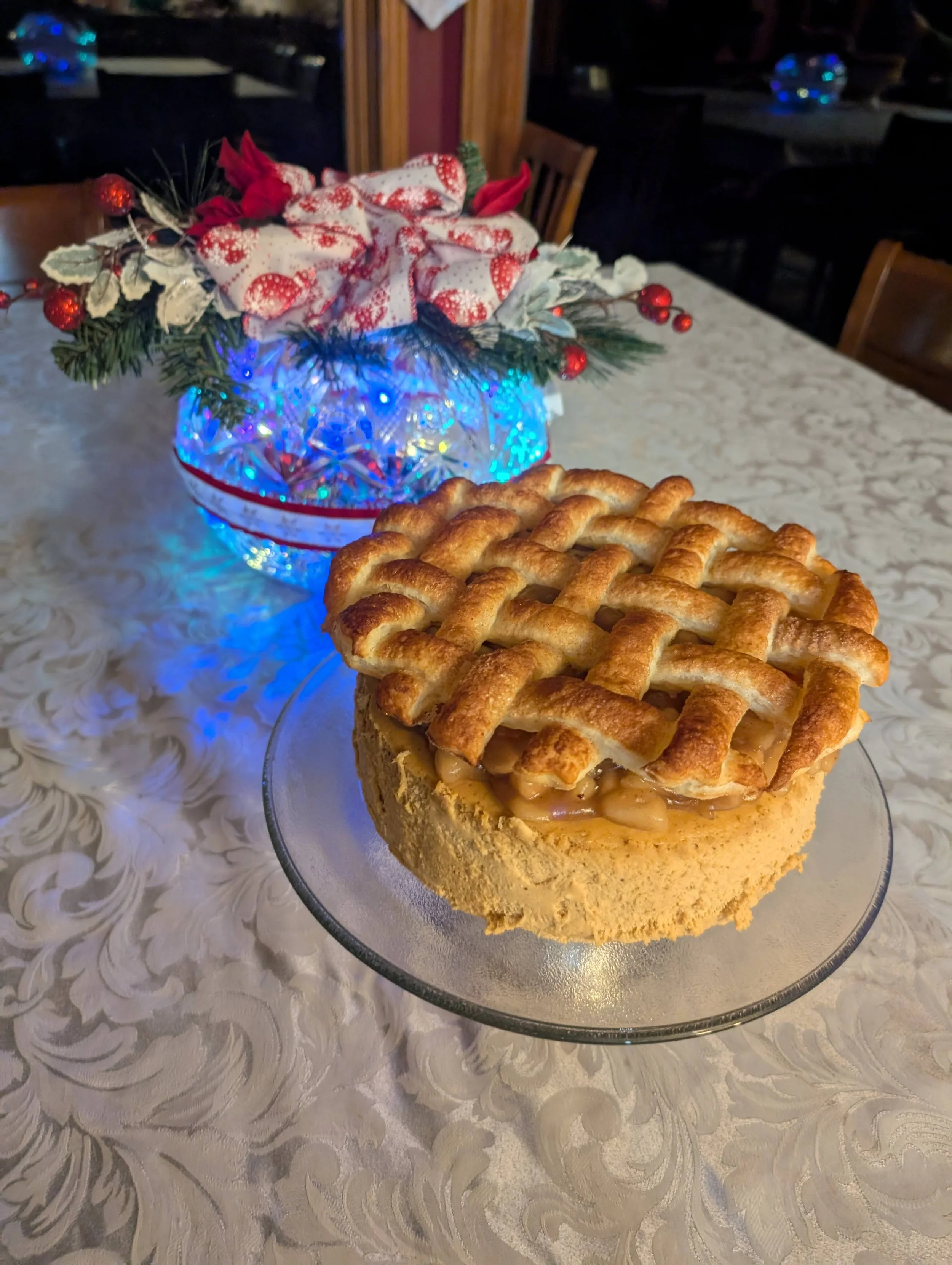 A apple pie with a lattice crust on a clear glass plate, placed on a white decorative tablecloth. In the background, a holiday-themed centerpiece with a bow, poinsettias, and red berries, illuminated with blue string lights.