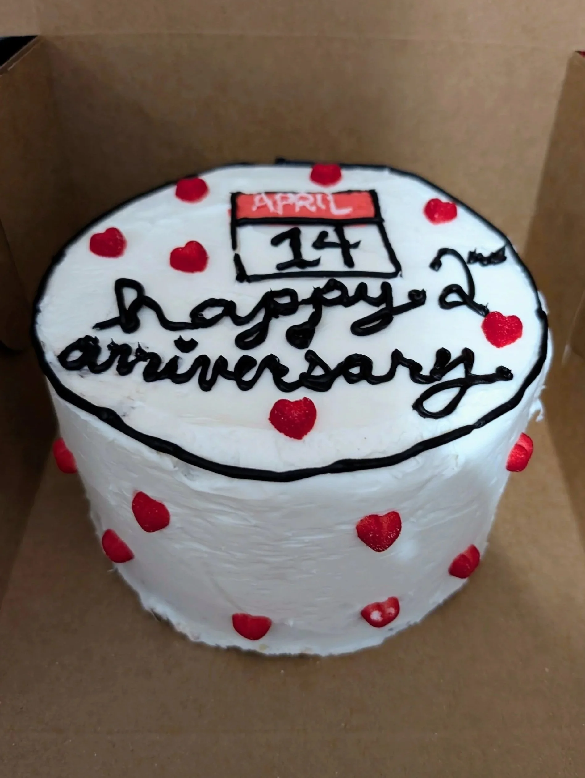 Heart-shaped birthday cake with white frosting, red heart decorations, and black writing that says 'happy 2nd anniversary' with a small calendar showing April 14.