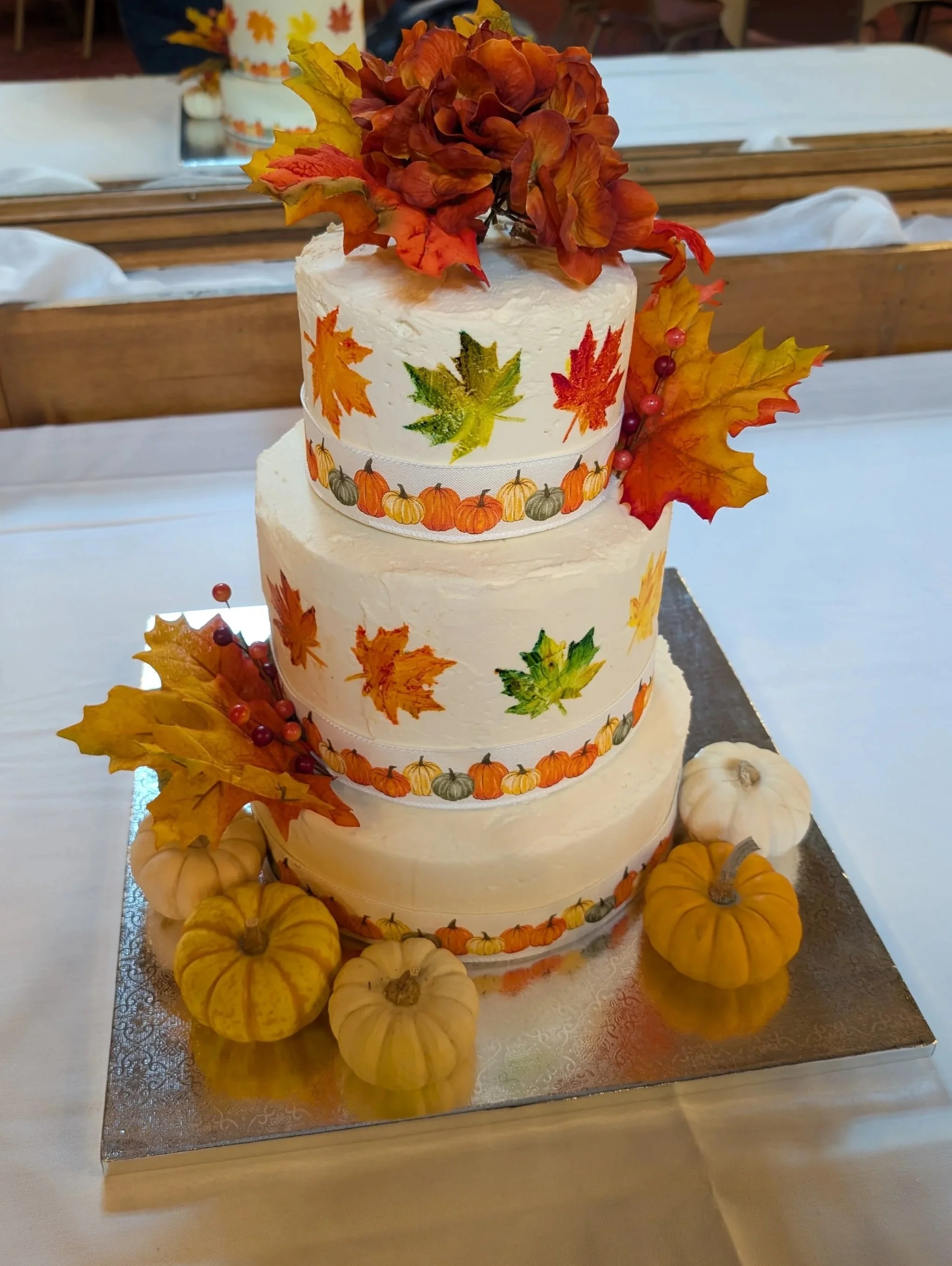 A three-tiered fall-themed cake decorated with autumn leaves, small pumpkins, and berries, surrounded by pumpkins on a silver board.