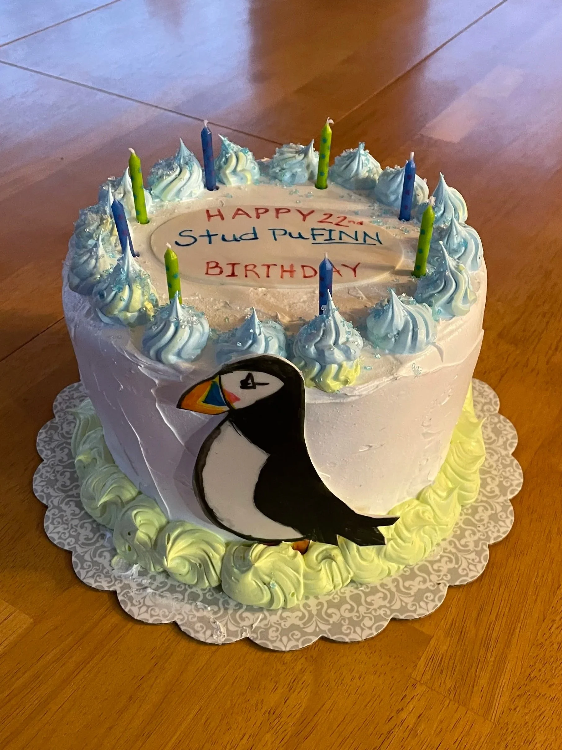 Birthday cake decorated with blue and white frosting, colorful candles, a penguin illustration on the side, and a message reading "HAPPY BIRTHDAY" 22nd Stud PUFINN  on top.