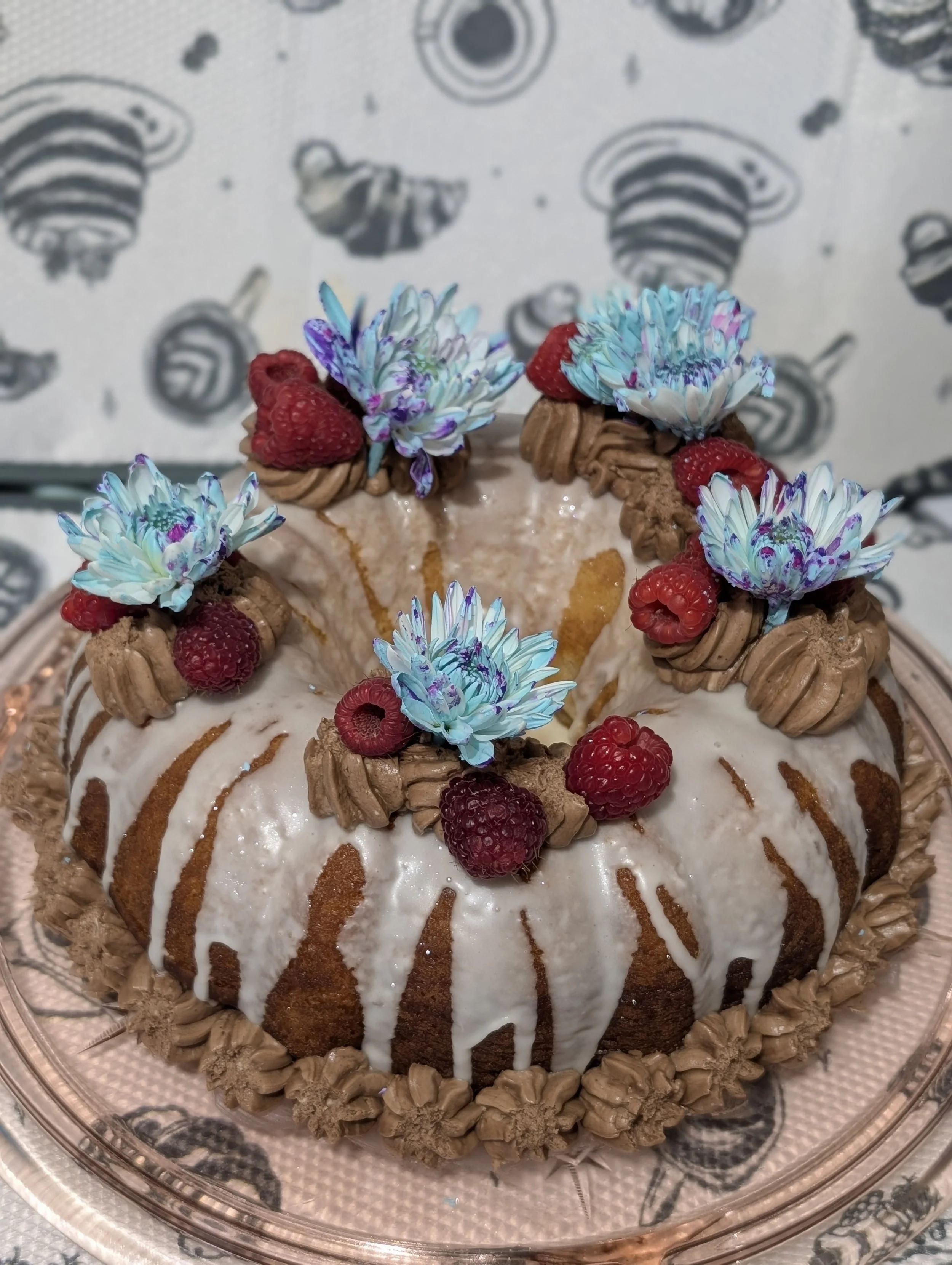 A bundt cake decorated with white icing, chocolate frosting, raspberries, and blue flowers, placed on a decorative plate.