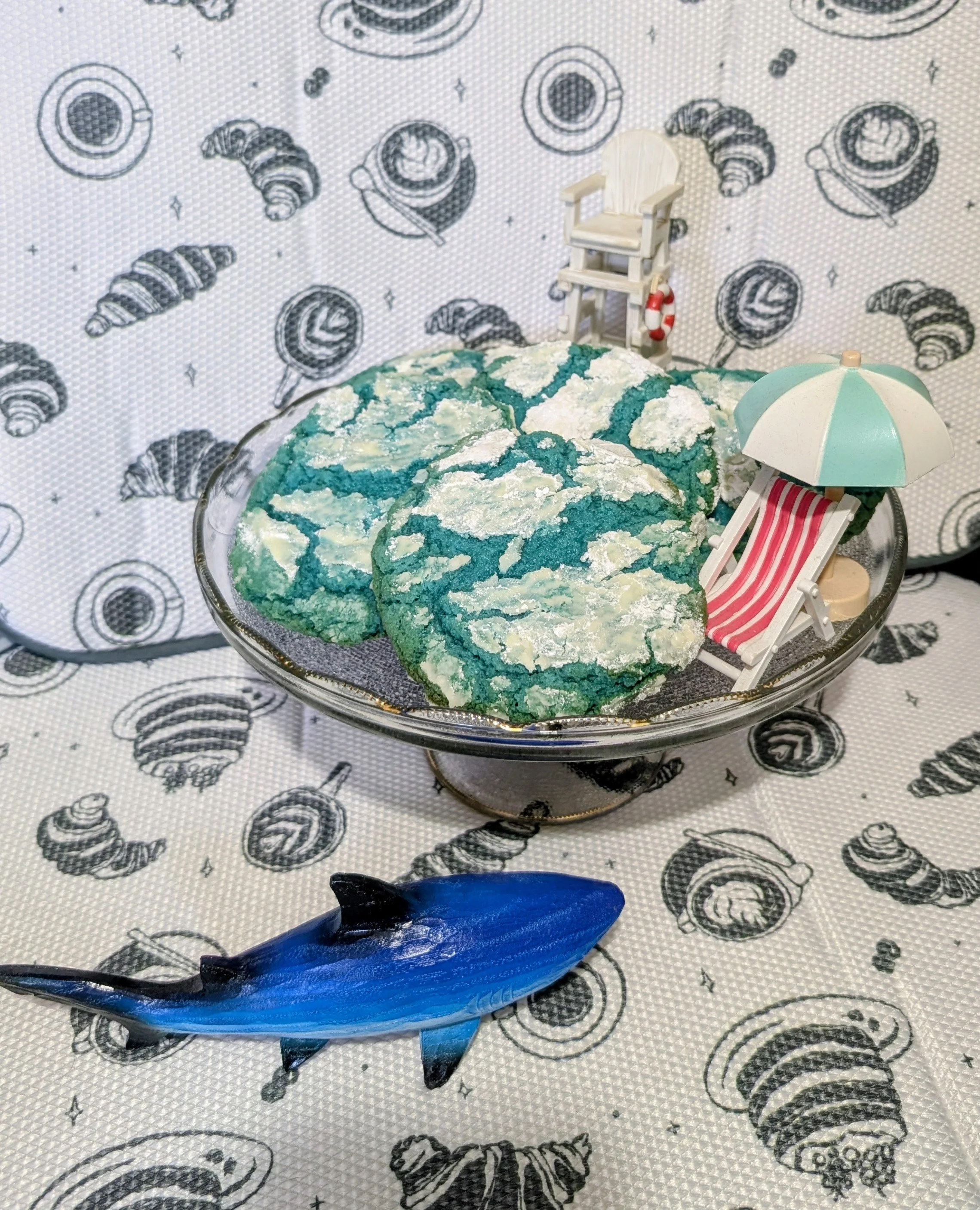 A glass cake stand with blue and white cracked cookies, mini beach chairs with umbrellas, a blue fish, and a shell-patterned background.