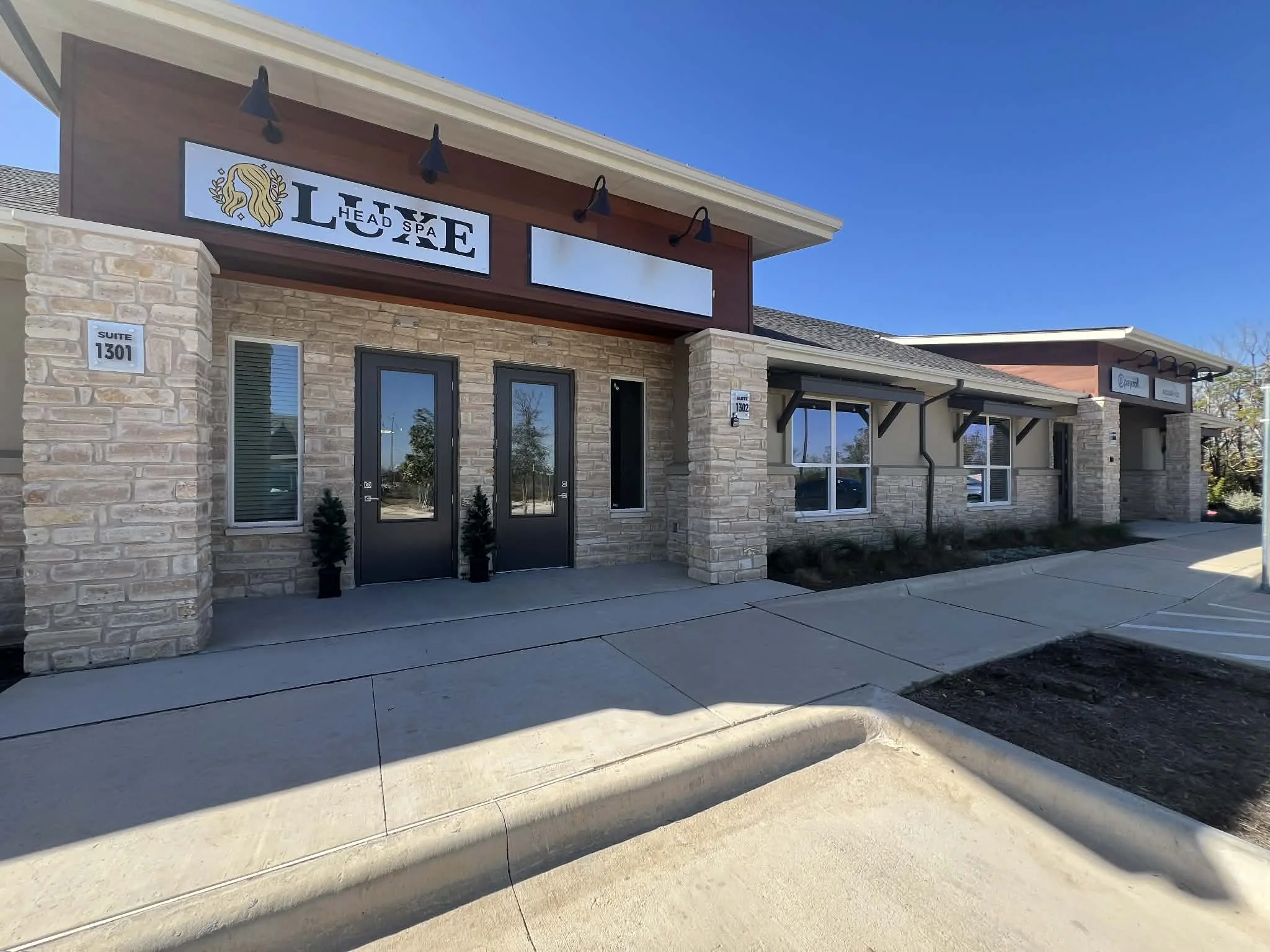 Exterior view of a modern stone and brick storefront with signs for LUXE Head Spa, and other businesses, under a clear blue sky.