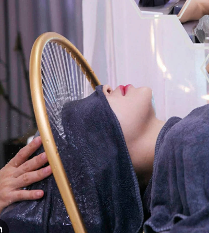 A person lying down with a dark hoodie pulled up over their head, resting their head on a wooden chair, and facing upward. Their face is visible in profile, with red lipstick, and water is splashed or dripping near their face.