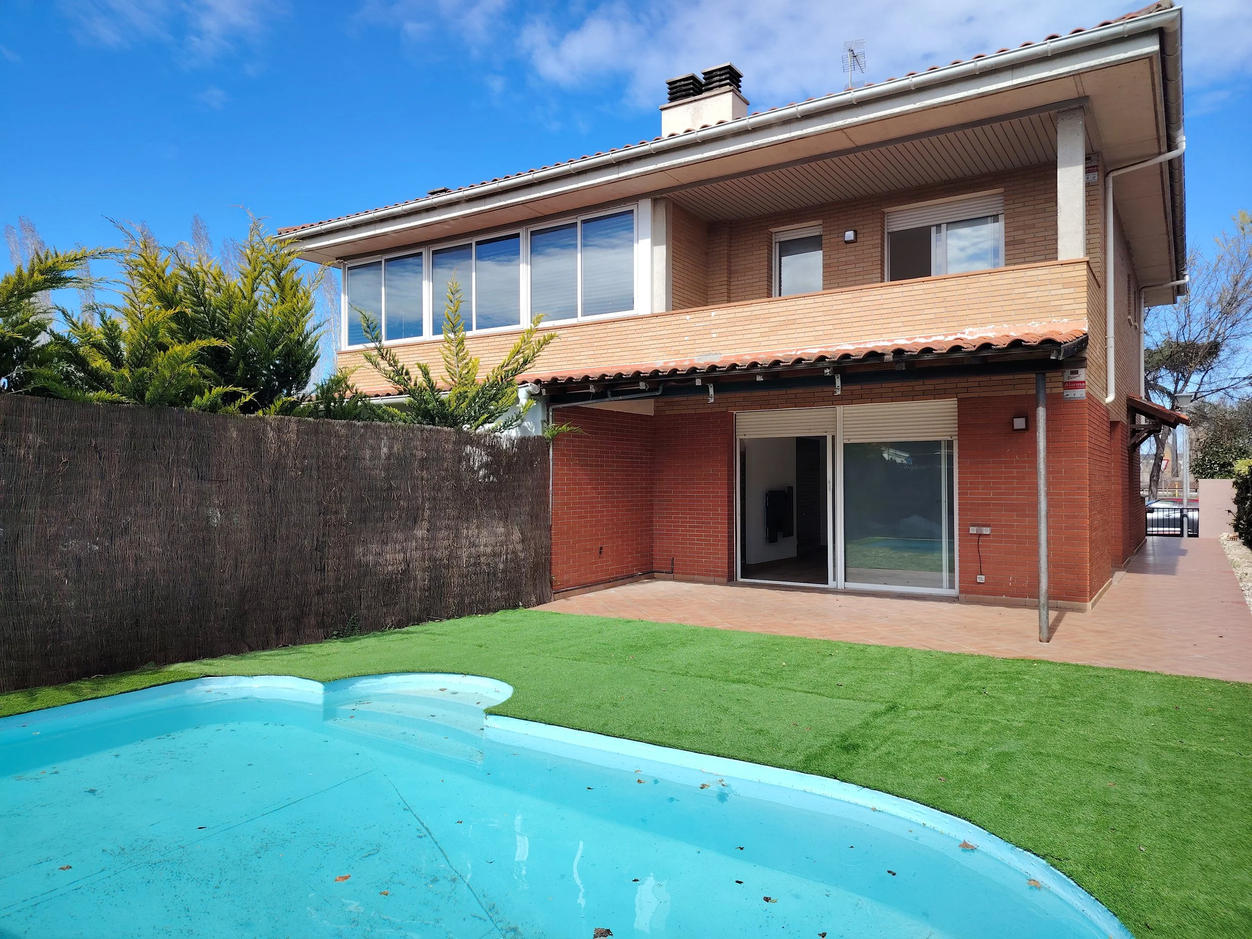 Casa de dos pisos con balcón, ventanas grandes, patio con césped y piscina azul, cielo azul y árboles en el fondo.