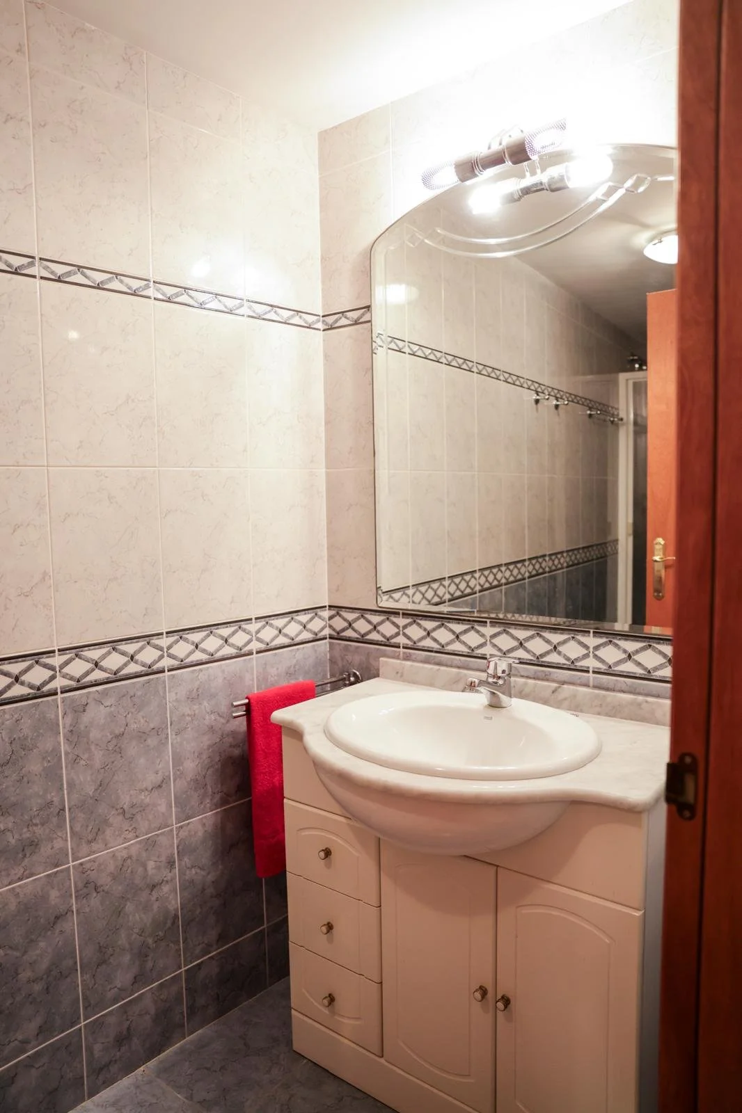 Un espejo grande refleja un lavabo con una cabina de ducha al fondo en un baño con paredes de azulejos beige con línea decorativa.