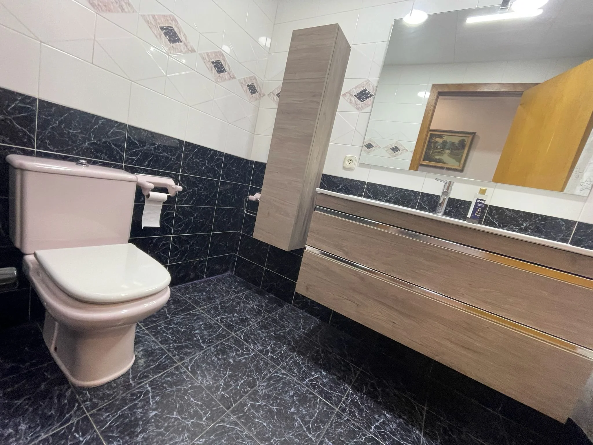 Baño con paredes de azulejo blanco con detalles en negro y tonos marrón, sanitario color rosa, lavabo con mueble de madera claro, espejo grande y un cuadro con paisaje en la pared.