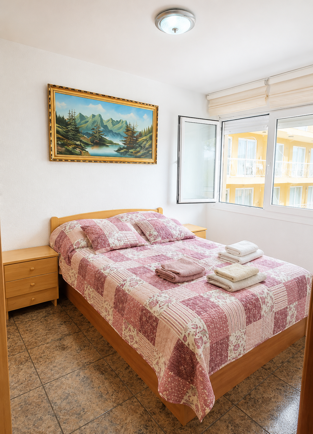Habitación con una cama con colcha y almohadas en tonos rosa, con toallas dobladas sobre ella, cerca de una ventana con vista a edificios amarillos con balcones, y un cuadro de paisaje montañoso y naturaleza en la pared.