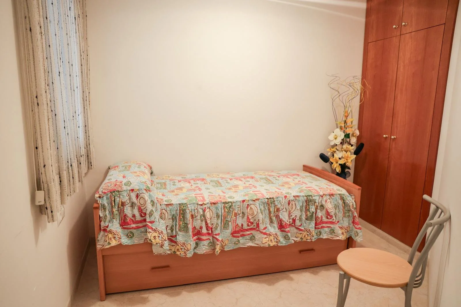 Habitación con cama individual, cortinas, armario de madera, florero con flores artificiales y silla de madera.