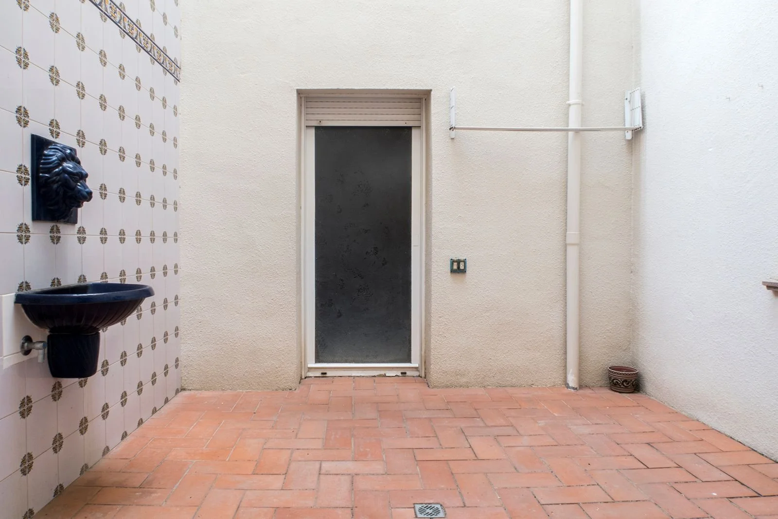 Patio interior con piso de ladrillos, pared blanca, puerta de vidrio y paredes decoradas con azulejos con patrón floral, lavabo de cerámica negra y un pequeño florero en la esquina derecha.
