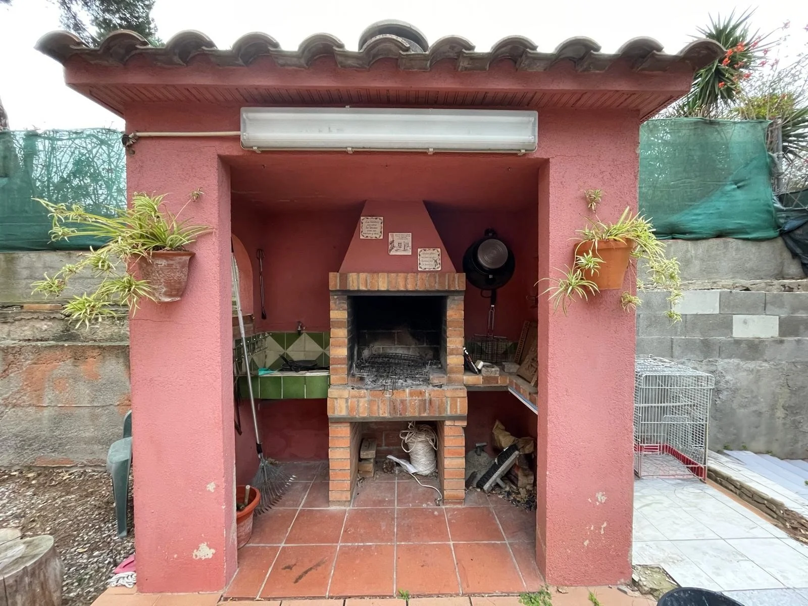 Mozuelo de ladrillos con estructura para barbacoa en el exterior, con plantas en macetas a los lados, utensilios de cocina y una jaula, en un fondo de muro de bloques de cemento y vegetación.