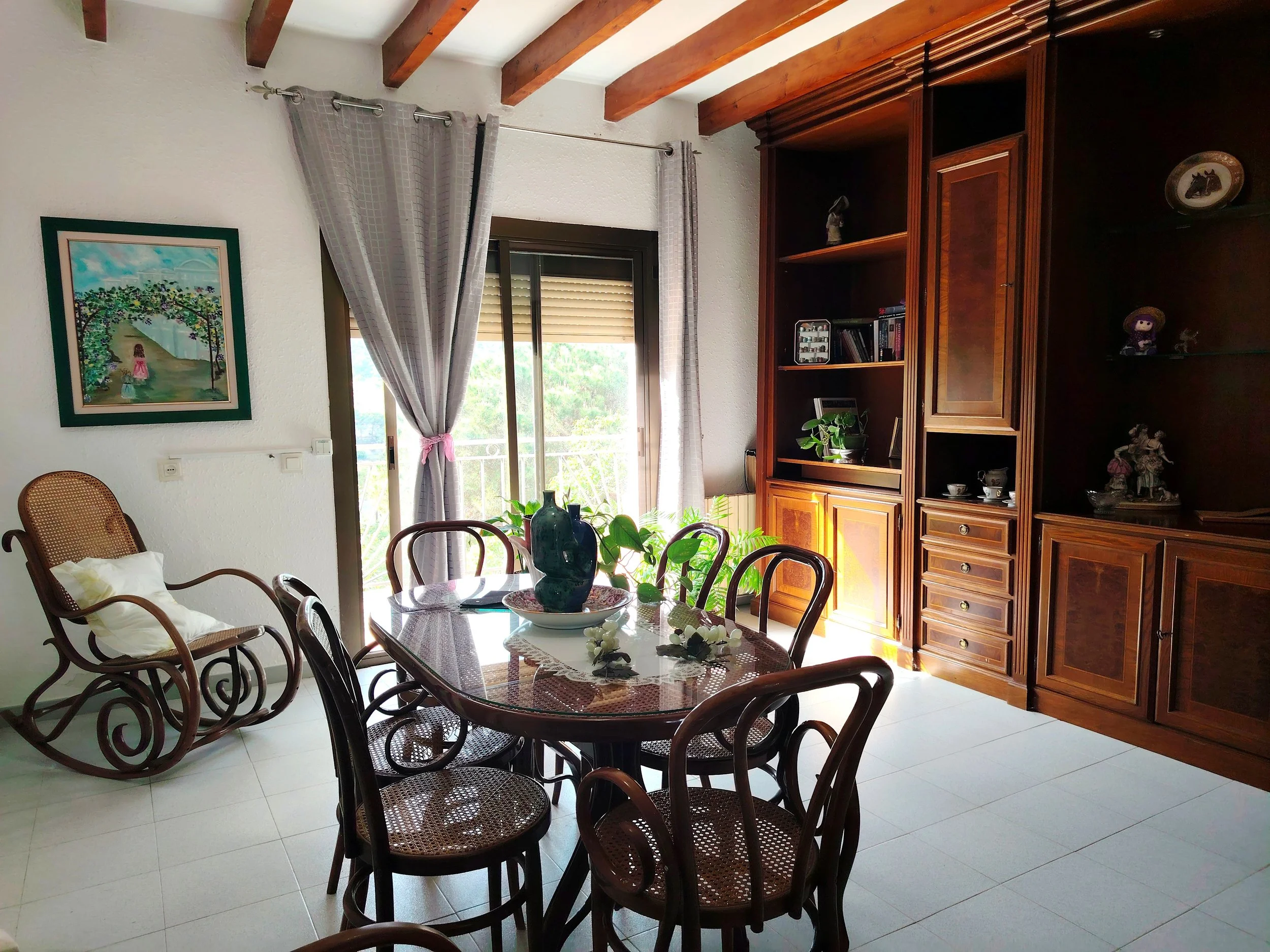 Vista del interior de un comedor con mesa de madera, sillas de mimbre, un armario grande de madera, cortinas gris claras y una pintura en la pared