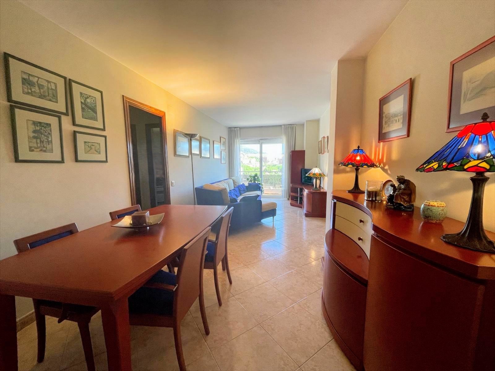 Sala de estar y comedor en un departamento con muebles de madera, cuadros en la pared, lámparas con pantallas coloridas y balcón con vista a la ciudad.