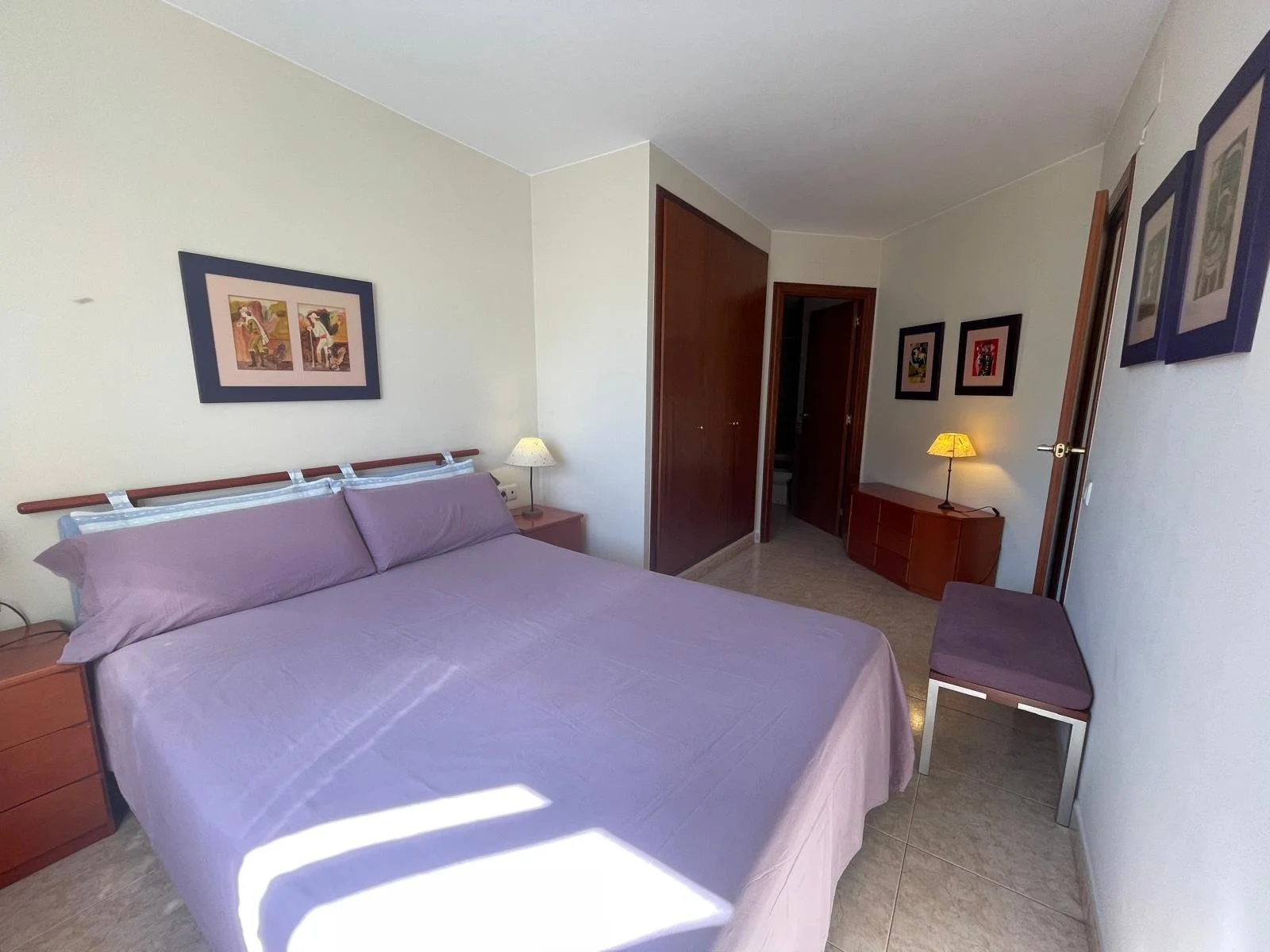 Habitación de hotel con cama grande, ropa de cama morada, dos mesas de noche con lámparas, cuadros en las paredes, armario de madera, y un asiento en la esquina.