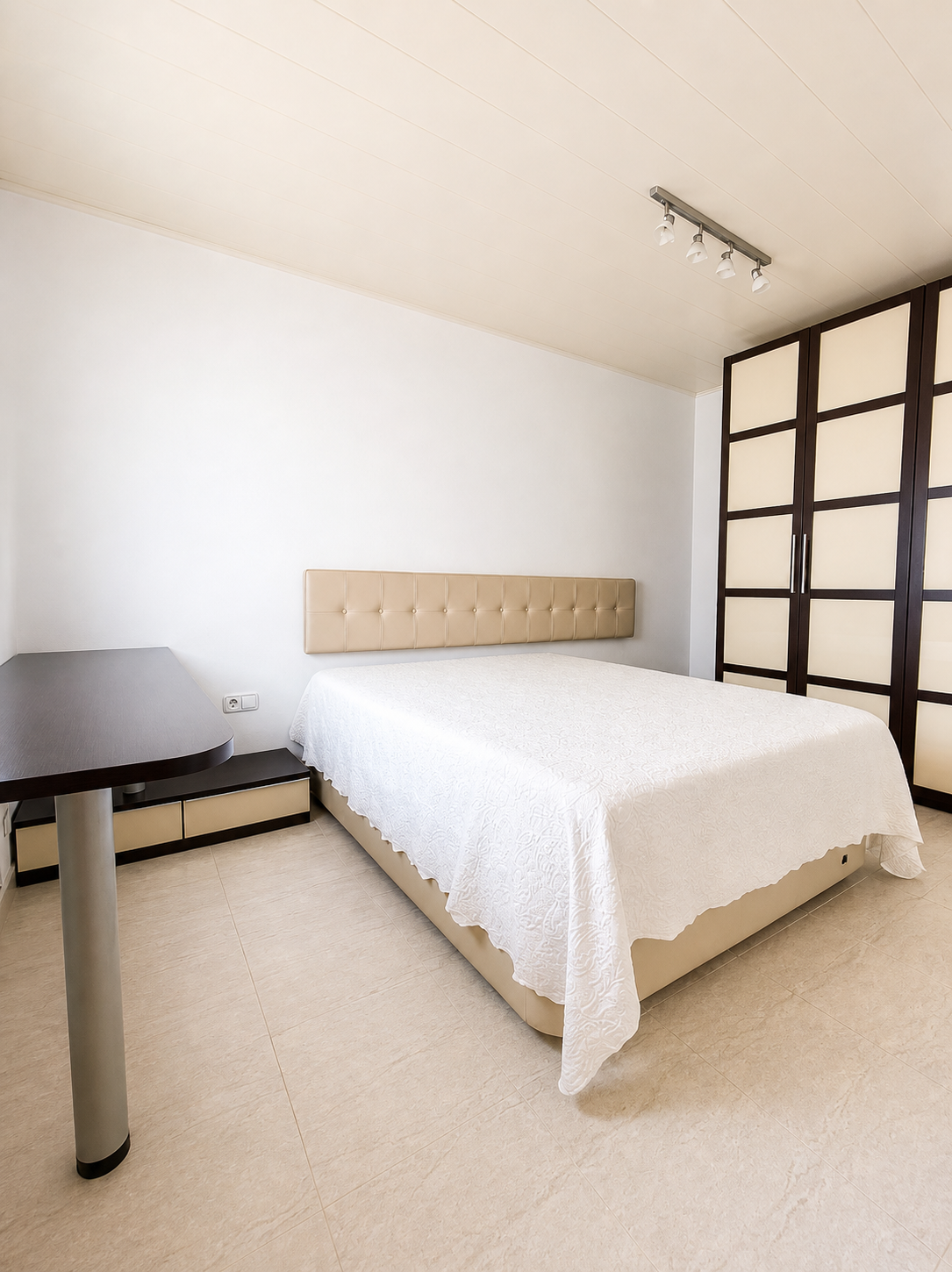 Habitación con cama, cabecera acolchada beige, armario grande de puertas oscuras y paneles claros, mesa de noche y pared blanca.