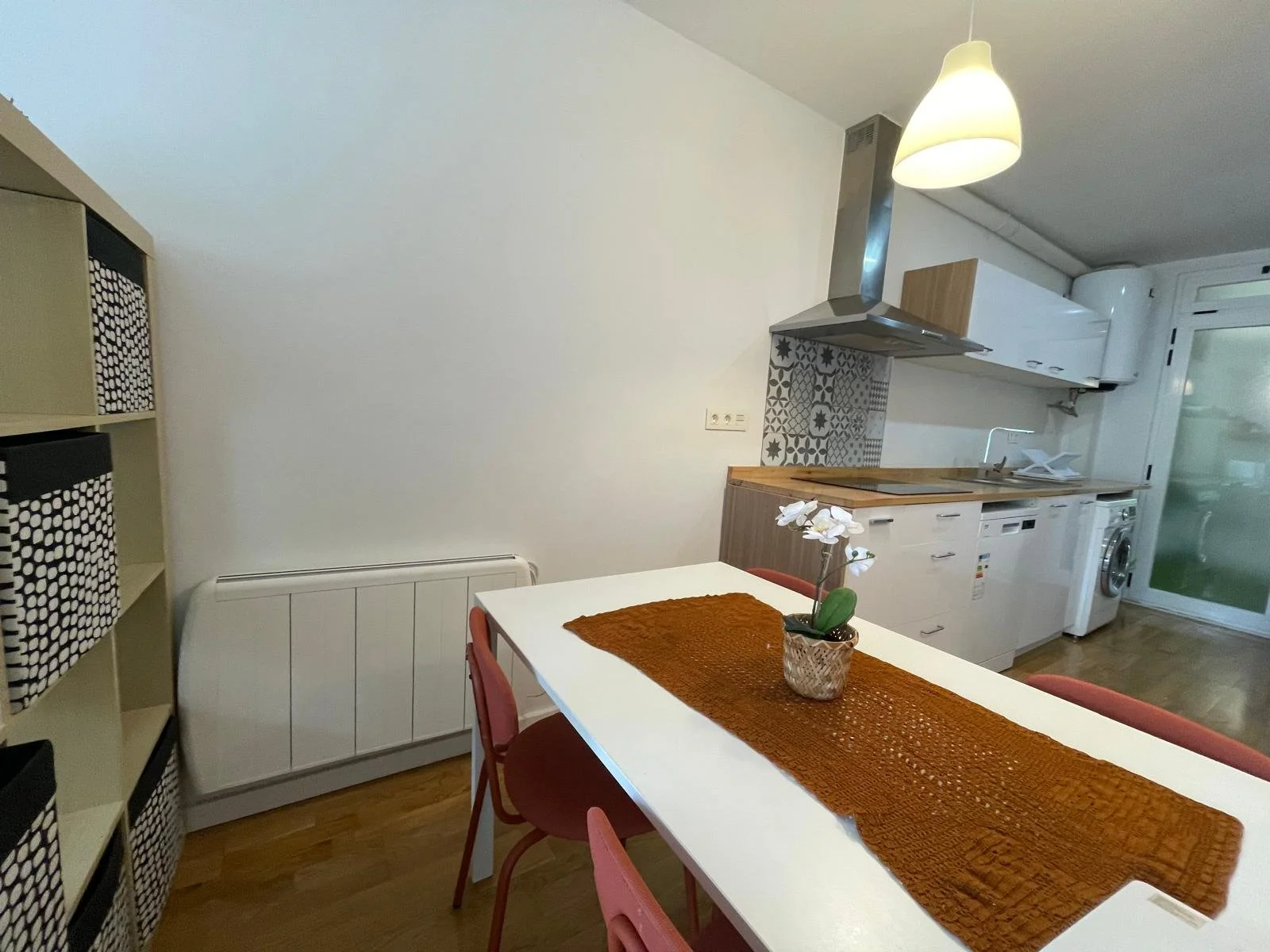 Cocina moderna con mesa de comedor blanca, decorada con un mantel naranja y una planta en una maceta de cristal.
