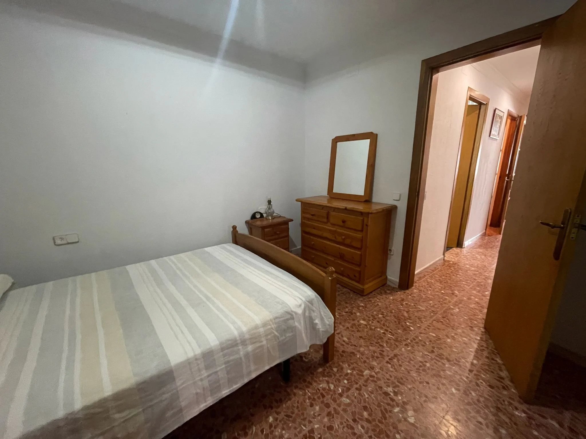 Habitación con cama, dos mesas de noche, un espejo y un armario de madera, en una casa con piso de baldosas y paredes blancas.