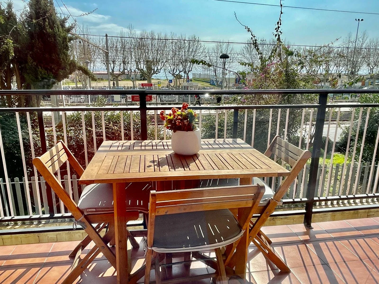 Vista de un balcón con una mesa de madera con una planta en un jarrón blanco en el centro, rodeada de cuatro sillas de madera. Al fondo se puede ver un parque con árboles, señalizaciones y una cancha deportiva bajo un cielo despejado.