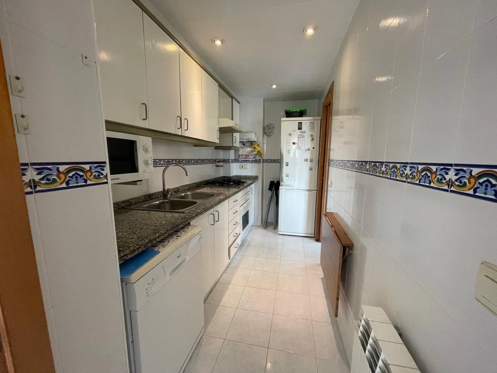 Cocina con gabinetes blancos, encimera de granito, lavabo doble, microondas, refrigerador, y piso de azulejos en tonos claros, decorada con borde de azulejos con diseño en azul, amarillo y verde.