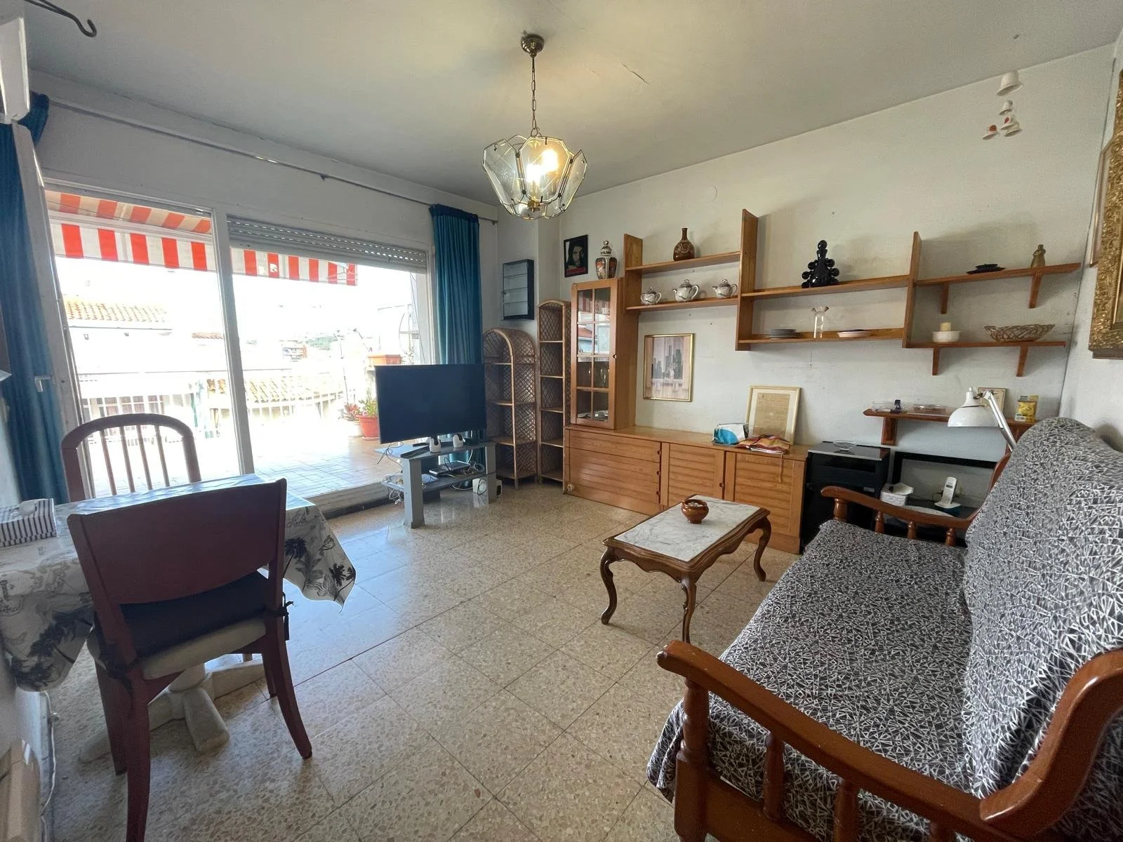 Sala de estar con sofá, mesa de café, estanterías con libros y objetos, televisor, silla, lámpara de escritorio, cuadros en las paredes y ventana con cortinas azules.