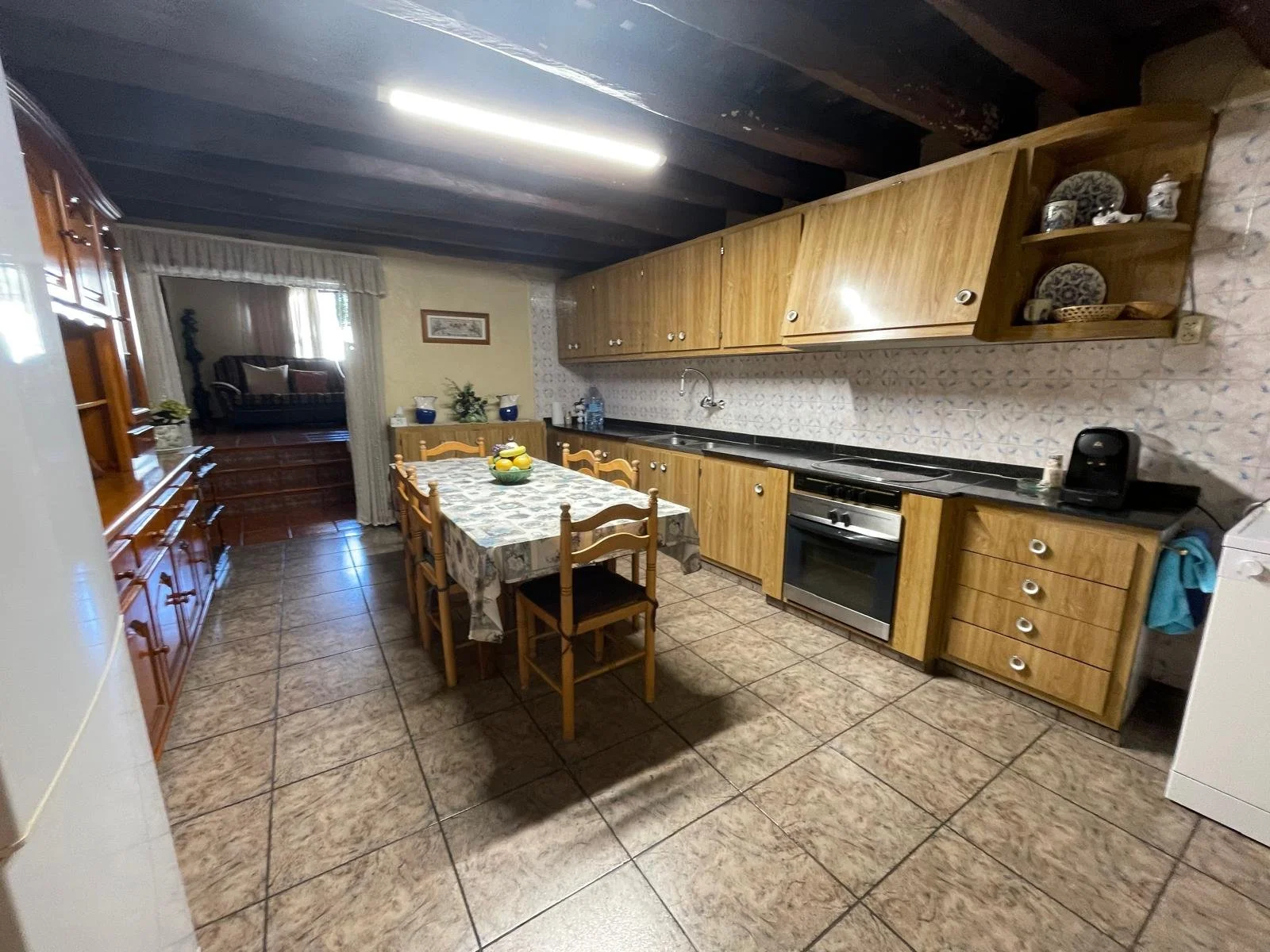 Cocina tradicional con gabinetes de madera y suelo de baldosas. Tiene comedor con mesa y sillas, y conecta a una sala de estar con sofá. Casa en Malgrat de mar.