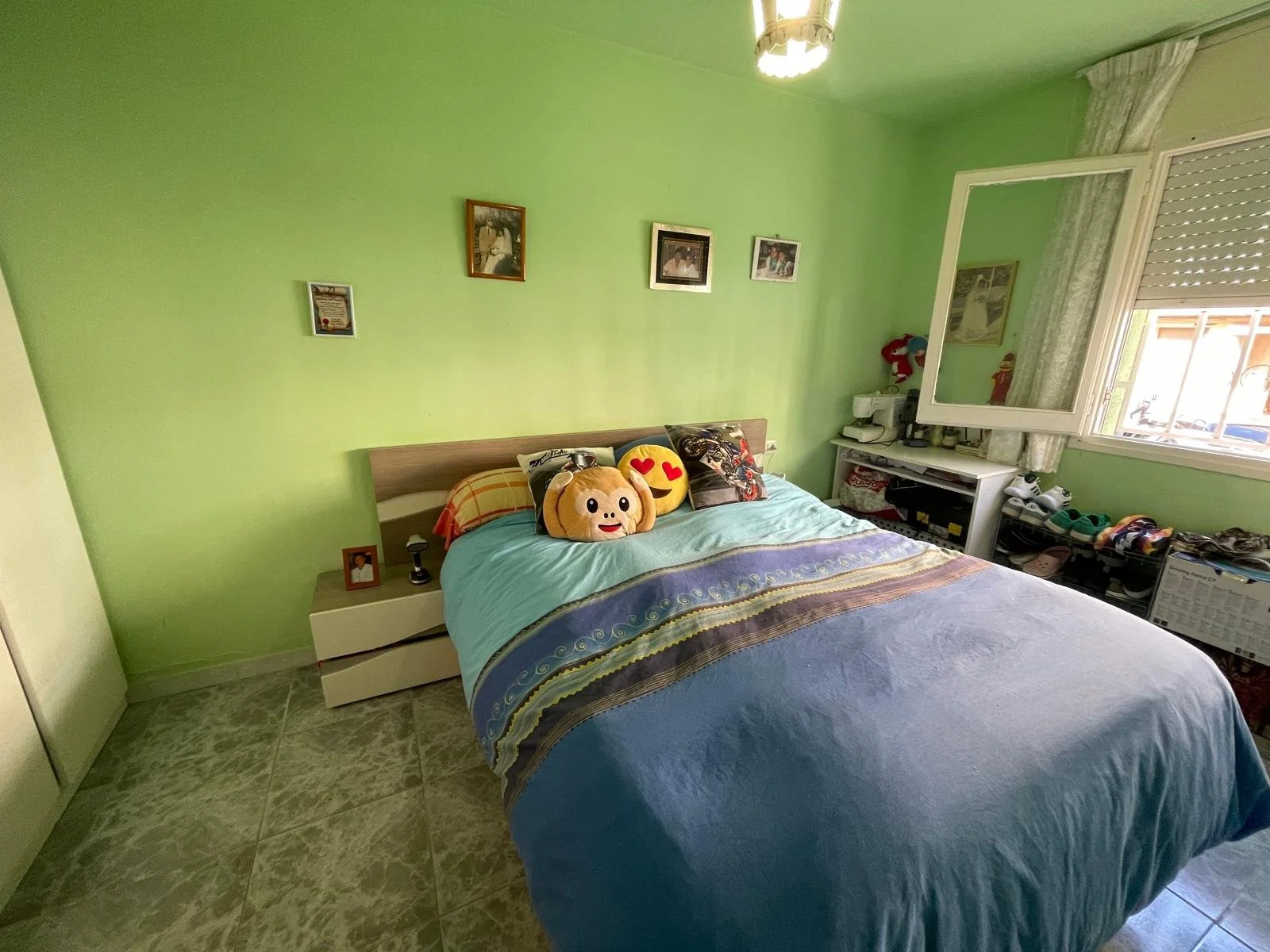 Habitación con paredes verdes, cama doblada con almohadas y peluches, ventana con cortinas y un espejo en la pared, espacio con zapatos y objetos diversos.