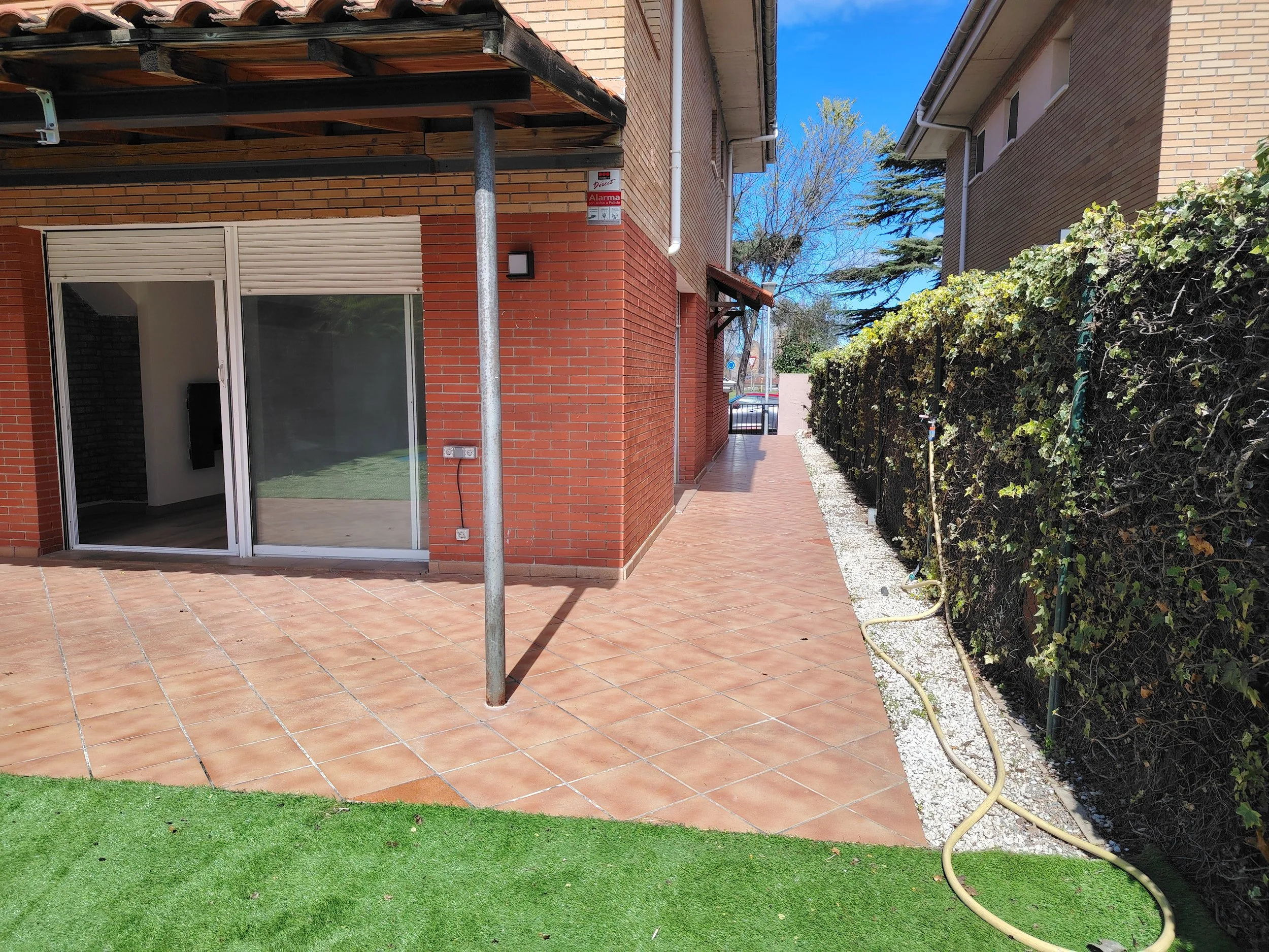 Patio exterior con piso de baldosas terracota, césped artificial, paredes de ladrillo y cerca de arbustos con una manguera en el suelo a un costado.
