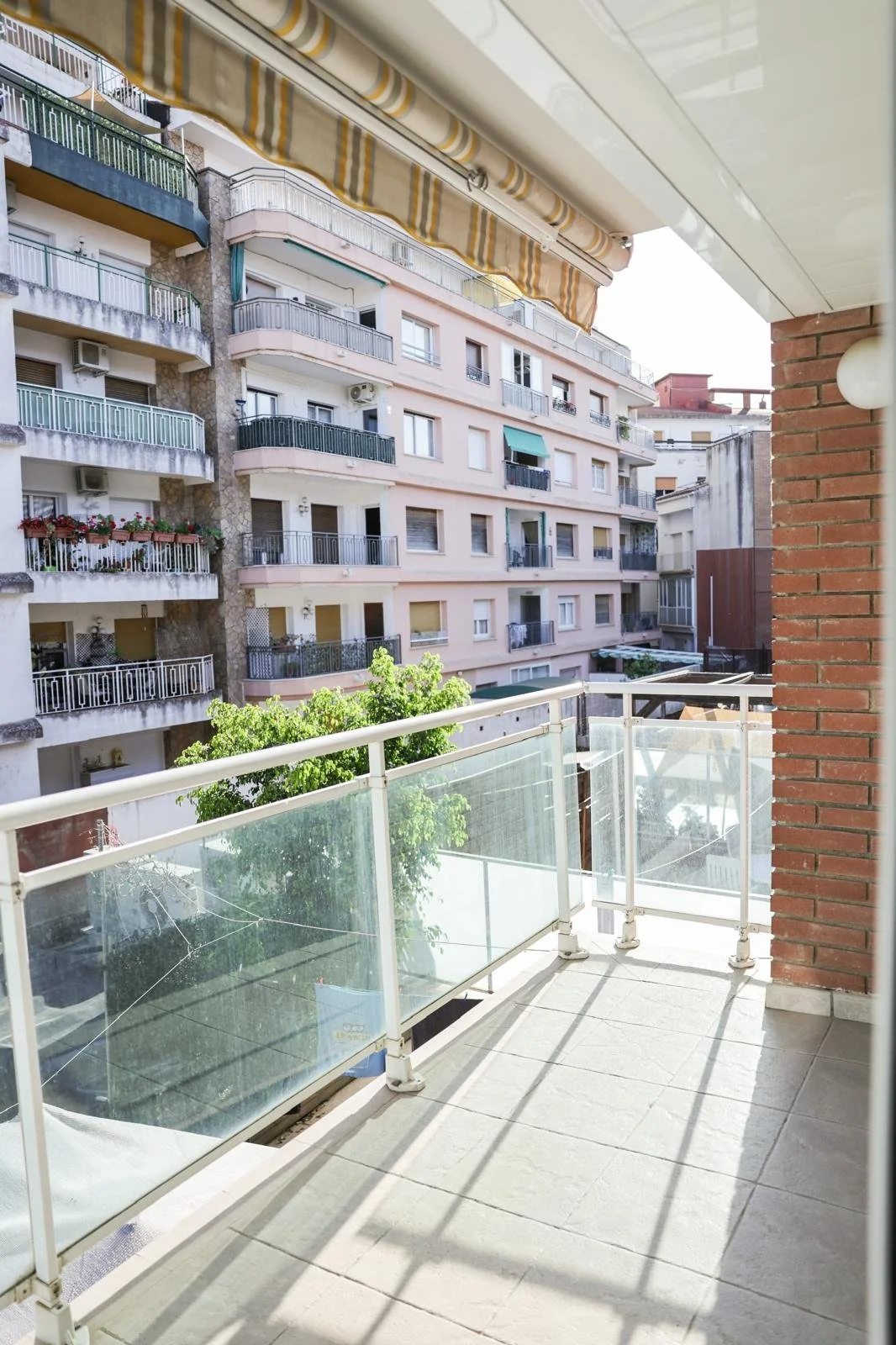 Vista de un balcón con barandal de vidrio, viendo edificios residenciales en una calle urbana, con un árbol y cielo despejado.