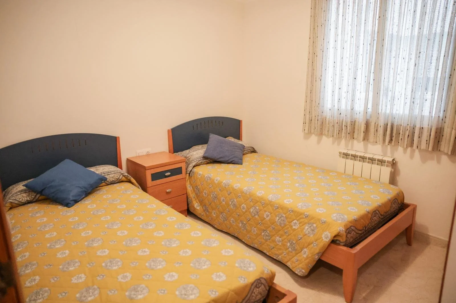 Habitación con dos camas doble con colchas amarillas y almohadas azules, una mesita de noche entre ellas, ventana con cortinas y radiador debajo.