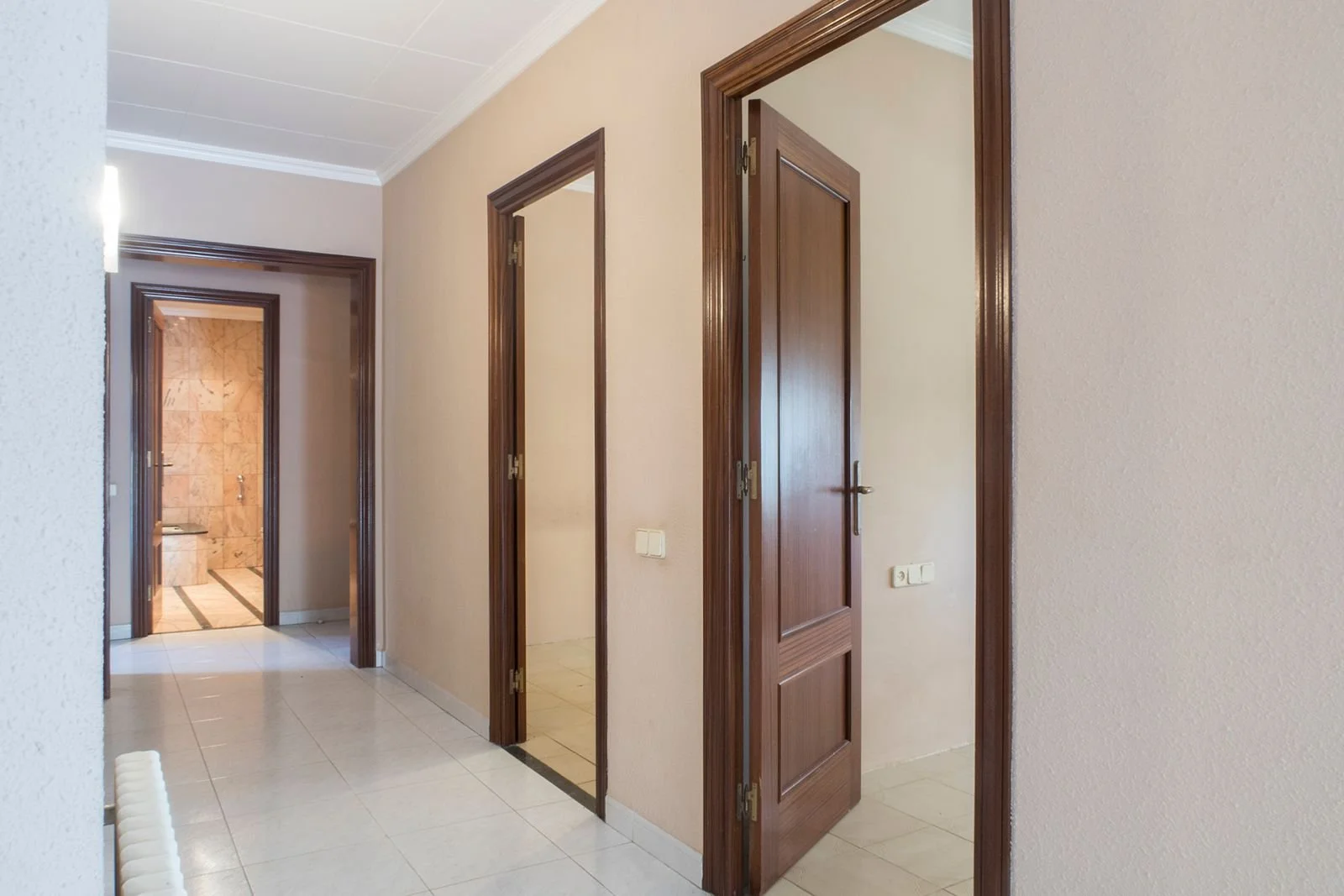 Pasillo de una casa con puertas de madera y paredes beige, un baño visible en el fondo.