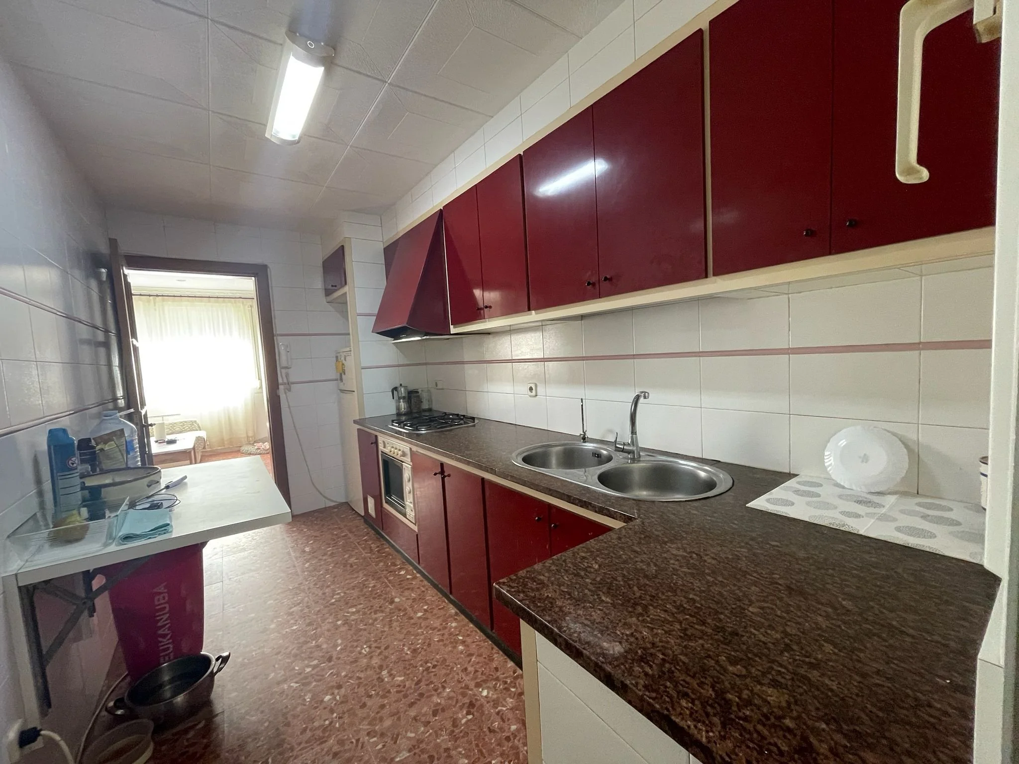 Vista de una cocina con gabinetes de color vino y encimeras de granito marrón oscuro. Tiene un fregadero doble y una estufa en la encimera. Hay una ventana y una puerta en el fondo con cortinas claras. En la mesa al frente hay utensilios y un rollo de papel aluminio. La pared está recubierta de azulejos blancos.
