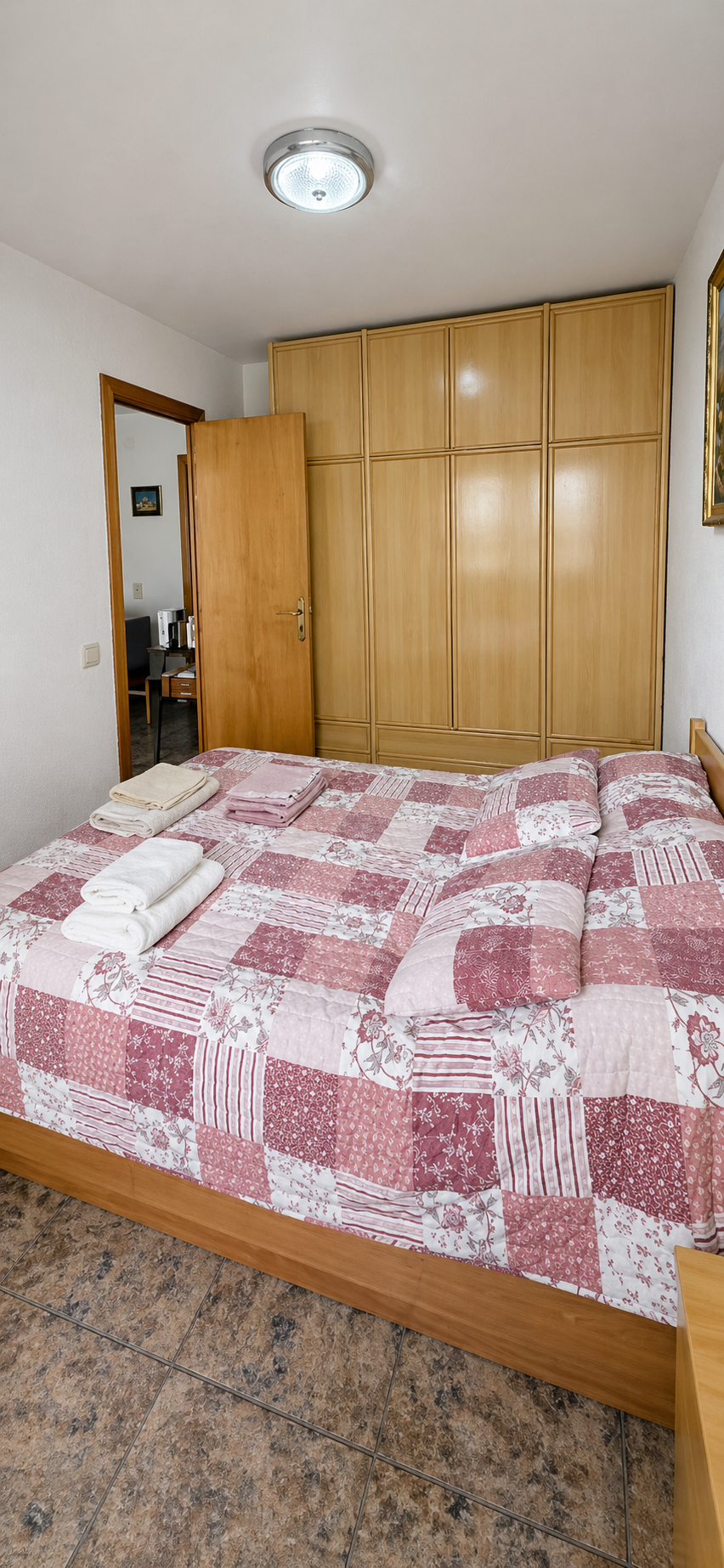 Habitación con cama de sábanas en tonos rosas y blancas, toallas dobladas en la cama, armario de madera y puerta abierta que muestra otra habitación con un escritorio.