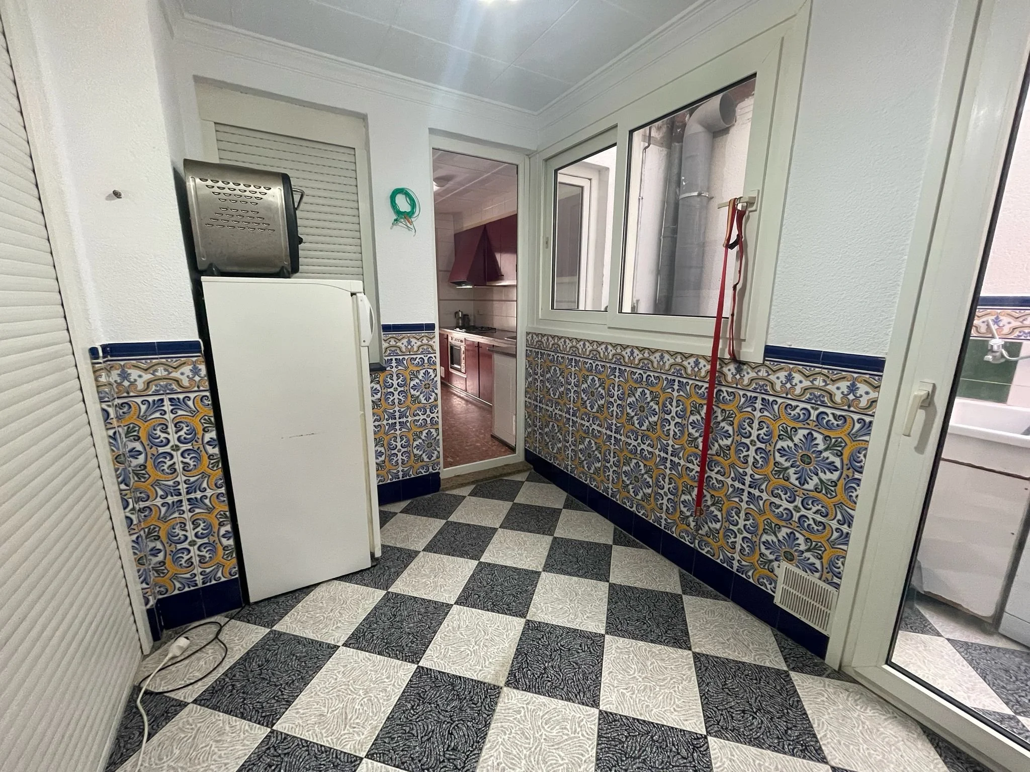 Una cocina con ventanas grandes, paredes blancas y azulejos decorativos en la parte inferior, piso de baldosas en patrón blanco y negro, y diversos objetos como una estufa, un refrigerador, mangueras y una escoba.