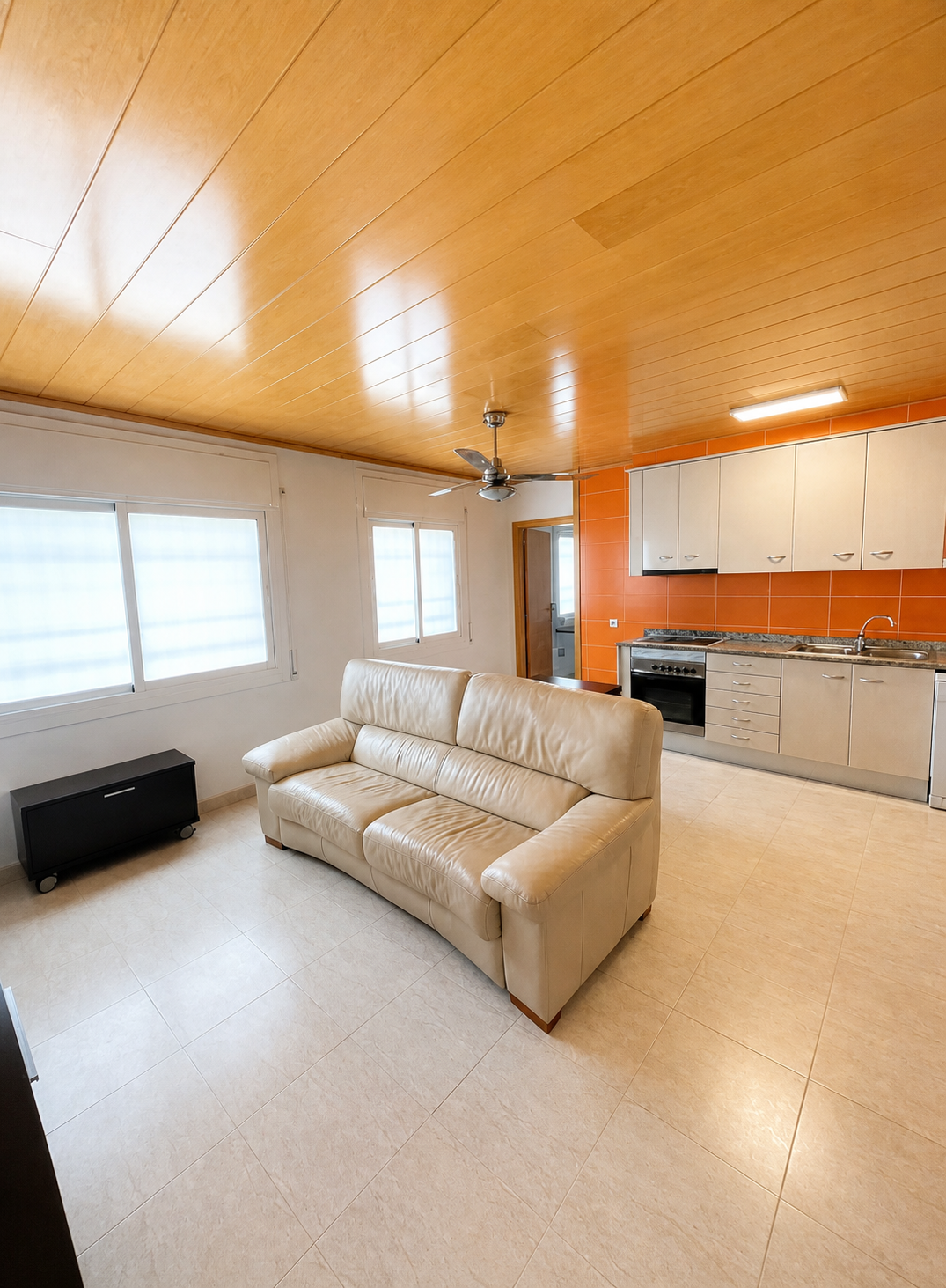Sala de estar con sofá beige, ventanas con cortinas blancas, cocina con gabinetes blancos y paredes de baldosas naranjas, techo de madera, ventilador y luz instalada