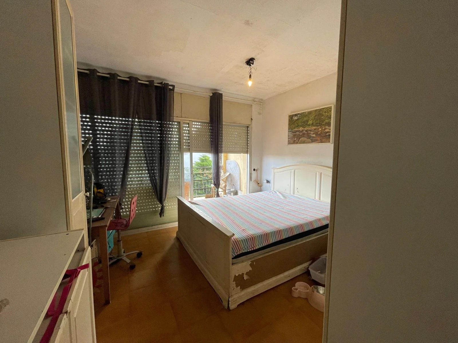 Habitación con cama de madera, sábanas a rayas y vista a un balcón con cortinas oscuras y ventana con persiana, paredes blancas, una foto en la pared y un escritorio con computador en la esquina izquierda.