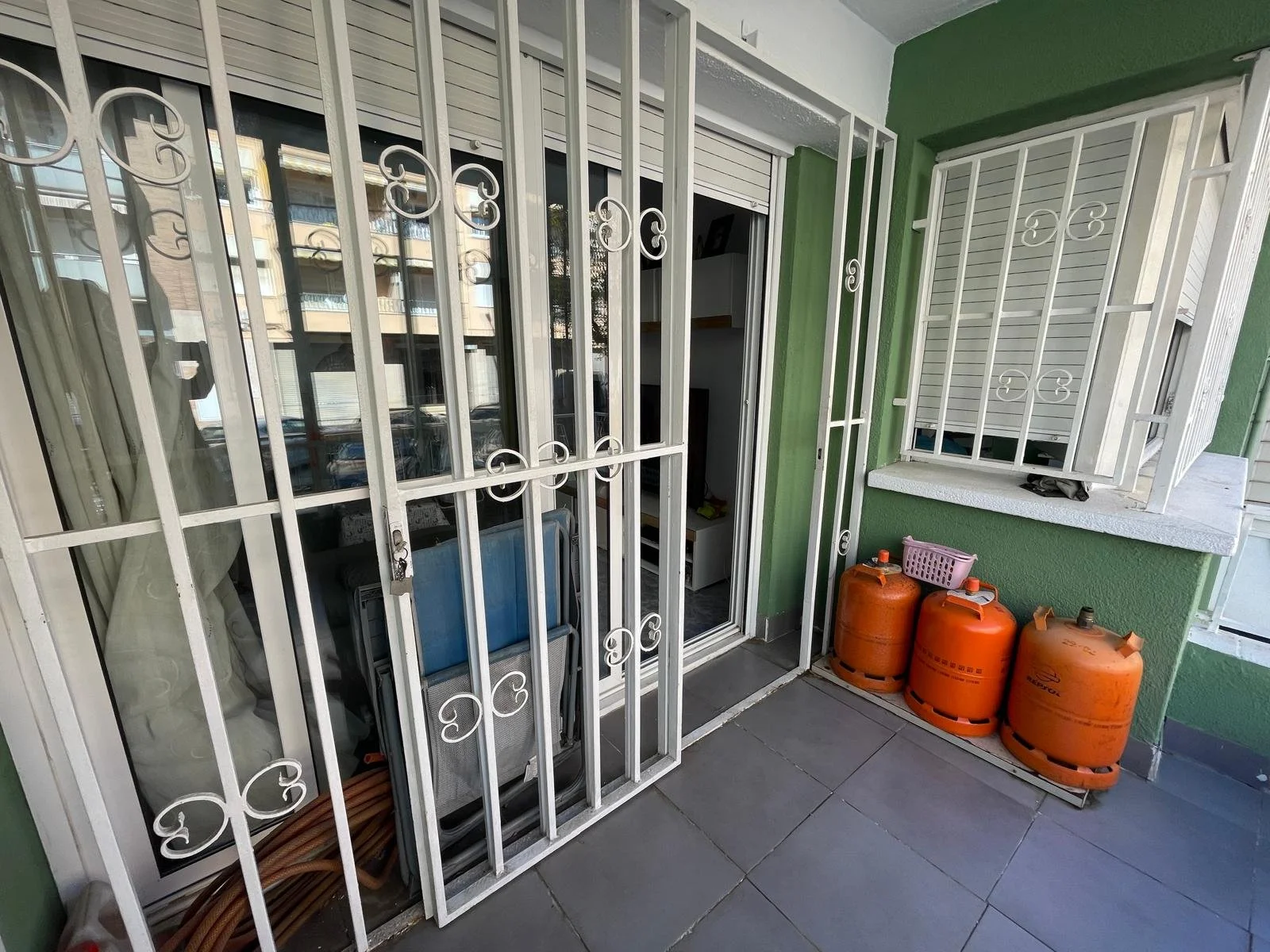 Balcón de apartamento con rejas blancas, dos cilindros de gas naranja, cesta morada, y vista a un interior y edificios exteriores.