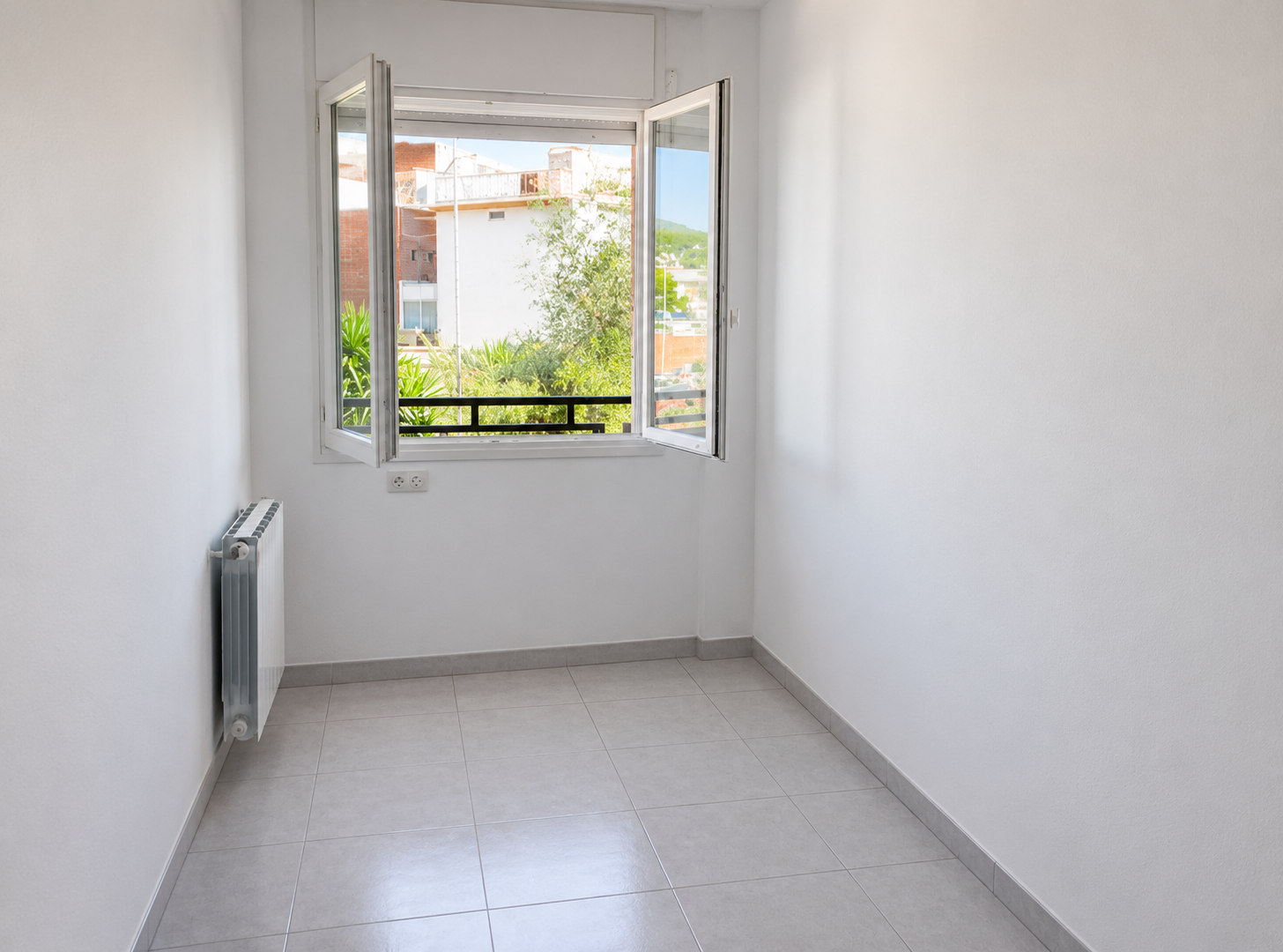 Habitación vacía con paredes blancas, piso de baldosas gris claro, ventana abierta con vista a árboles y edificios, y radiador debajo de la ventana.
