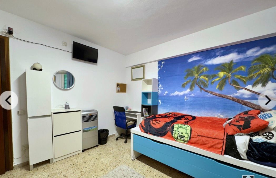 Habitación con pared decorada con mural de palmeras y playa, cama con sábanas de colores naranja y azul, escritorio con silla y televisor