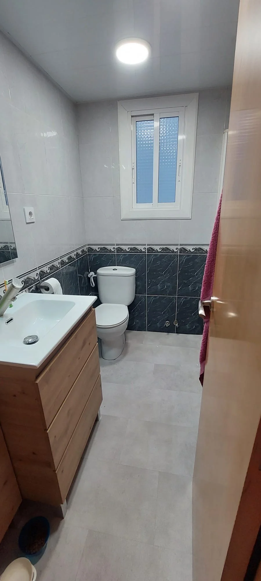 Baño con espejo, lavabo de madera, inodoro y ventana, paredes con azulejos blancos y azules, balón de agua sobre el inodoro, toalla rosa colgada, piso de baldosas claras.