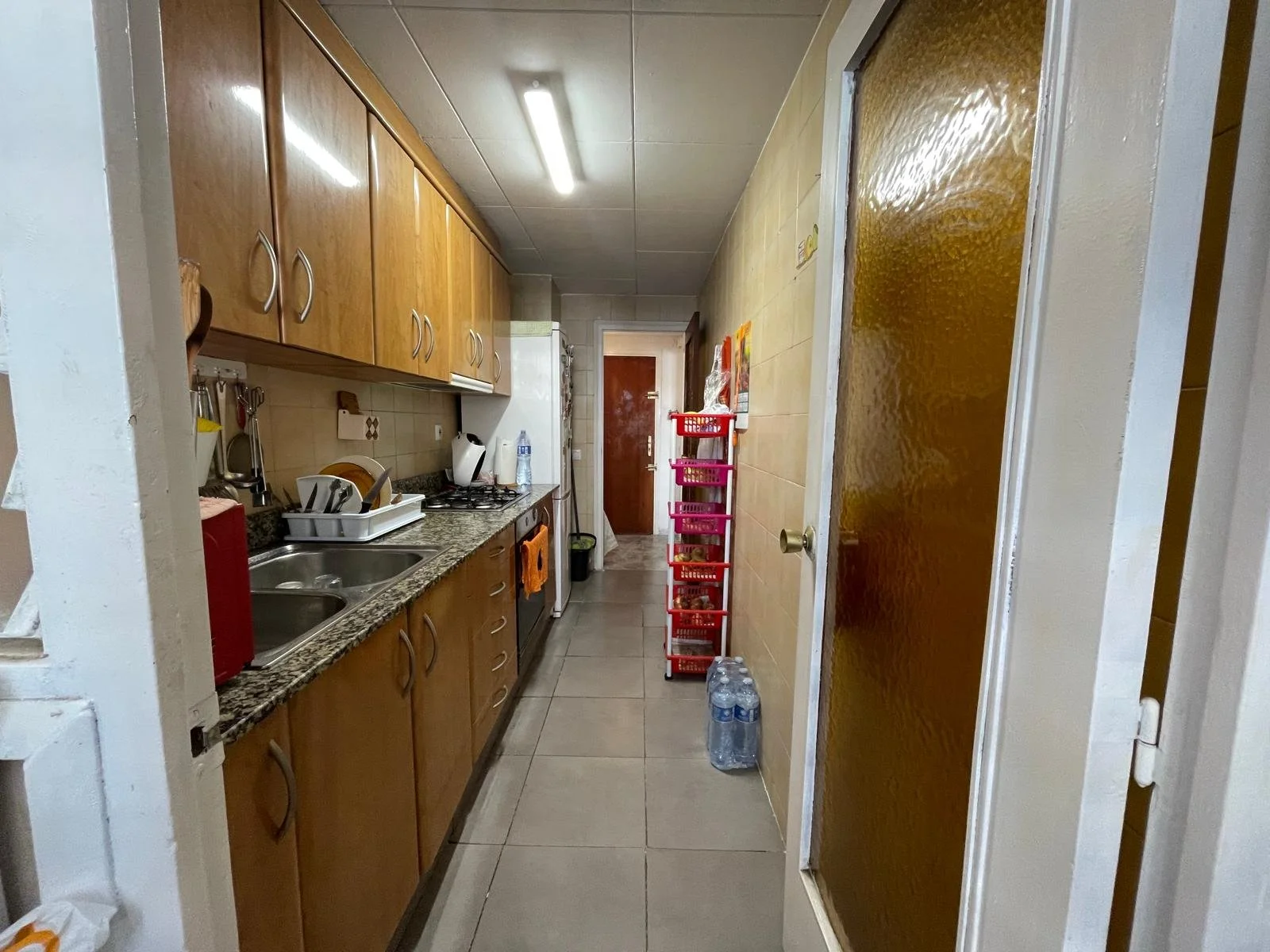 Cocina estrecha con gabinetes de madera, encimera de granito, fregadero doble, electrodomésticos y utensilios en la encimera, puerta con vidrio esmerilado a la derecha, pasillo con estantes de madera rosa, y botellas de agua en el suelo al final del pasillo.