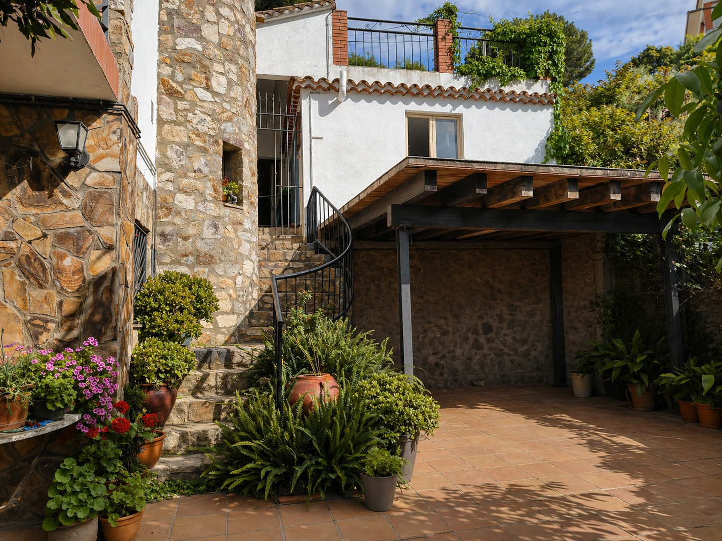Patio con plantas y paredes de piedra, con escaleras y un área techada de madera bajo una casa blanca, con cielo azul y árboles verdes.